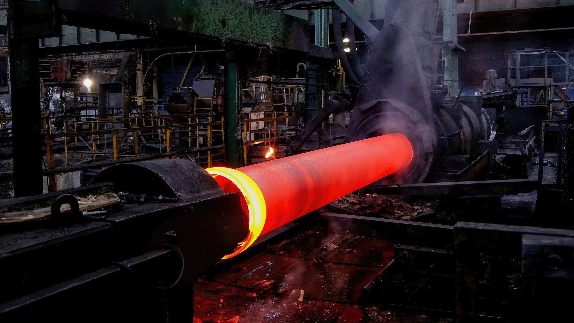 A glowing, red-hot steel pipe is processed by heavy industrial machinery in a factory setting.