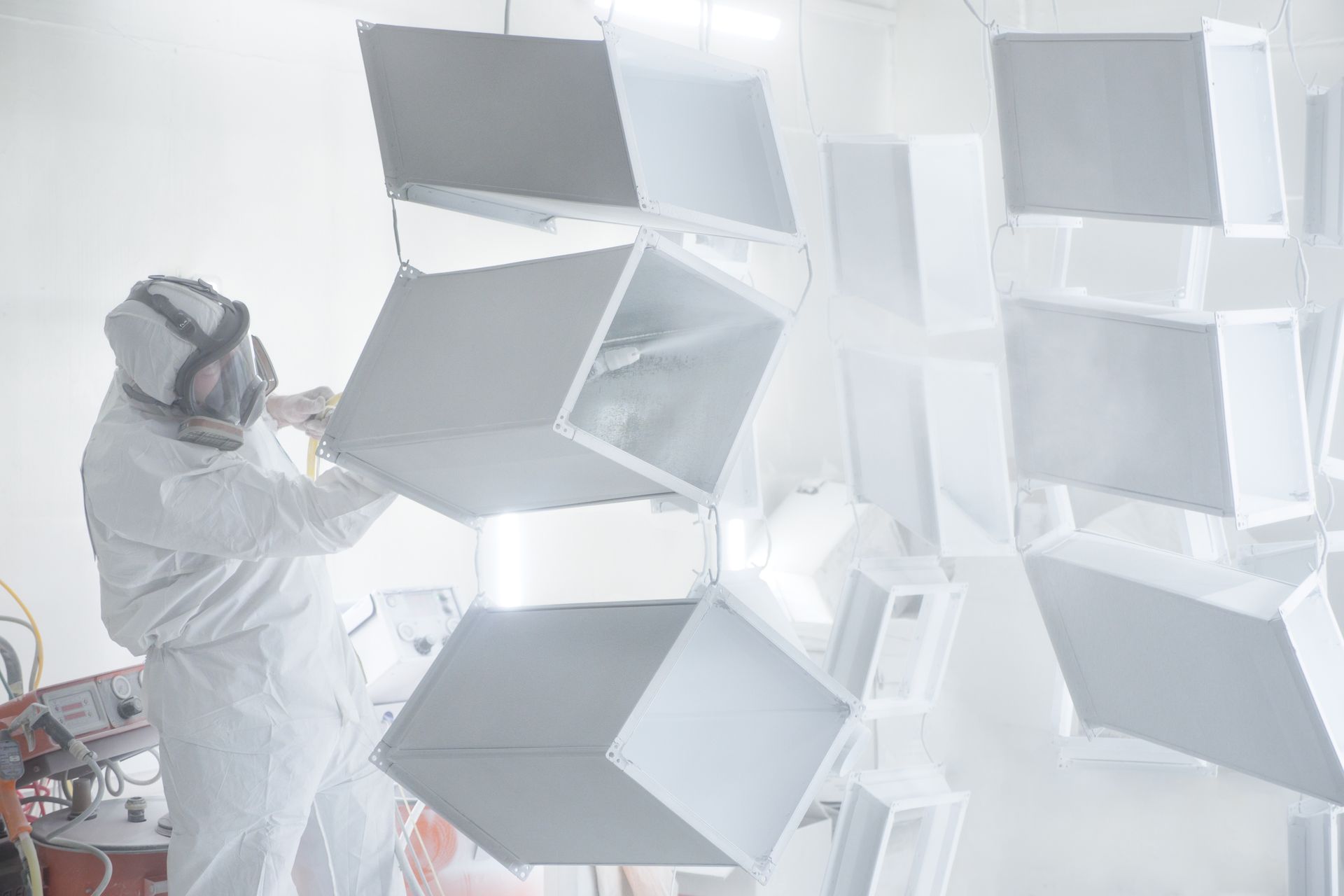 A person in protective gear sprays paint onto suspended white metal boxes in a workshop.