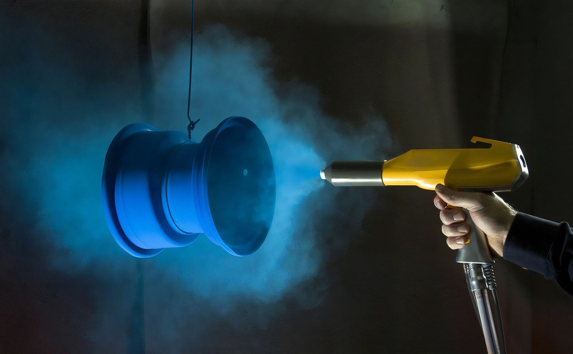 A hand using a yellow powder coating gun sprays blue powder onto a blue object.