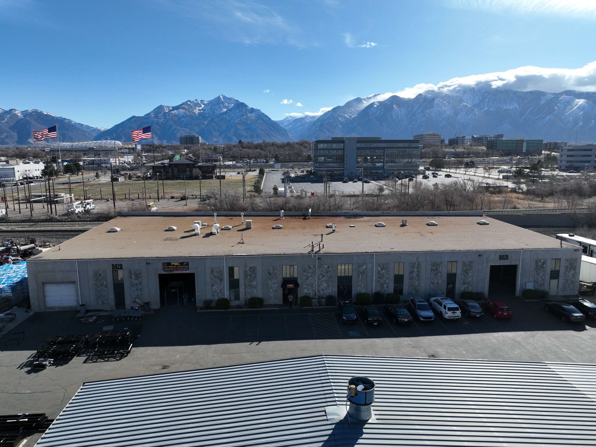 Large industrial building with parked cars, flags, and mountains in the background under a blue sky.