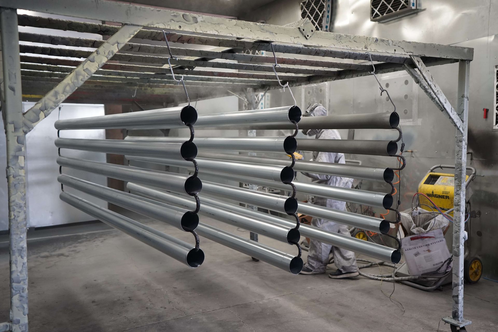 Metal pipes hanging in a spray paint booth, being coated with silver paint. Workers in protective gear are visible.