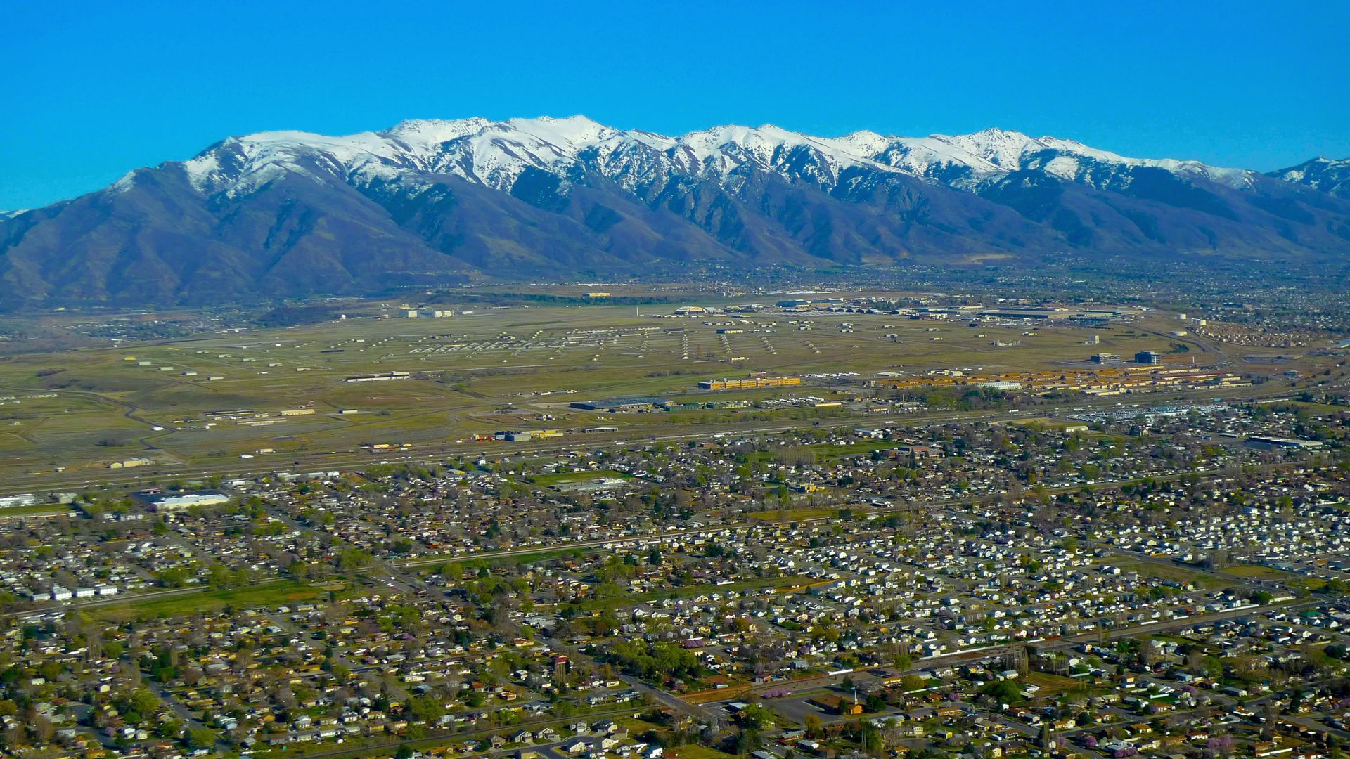 A sprawling town sits in a valley floor beneath a massive, snow-capped mountain range under a clear blue sky.