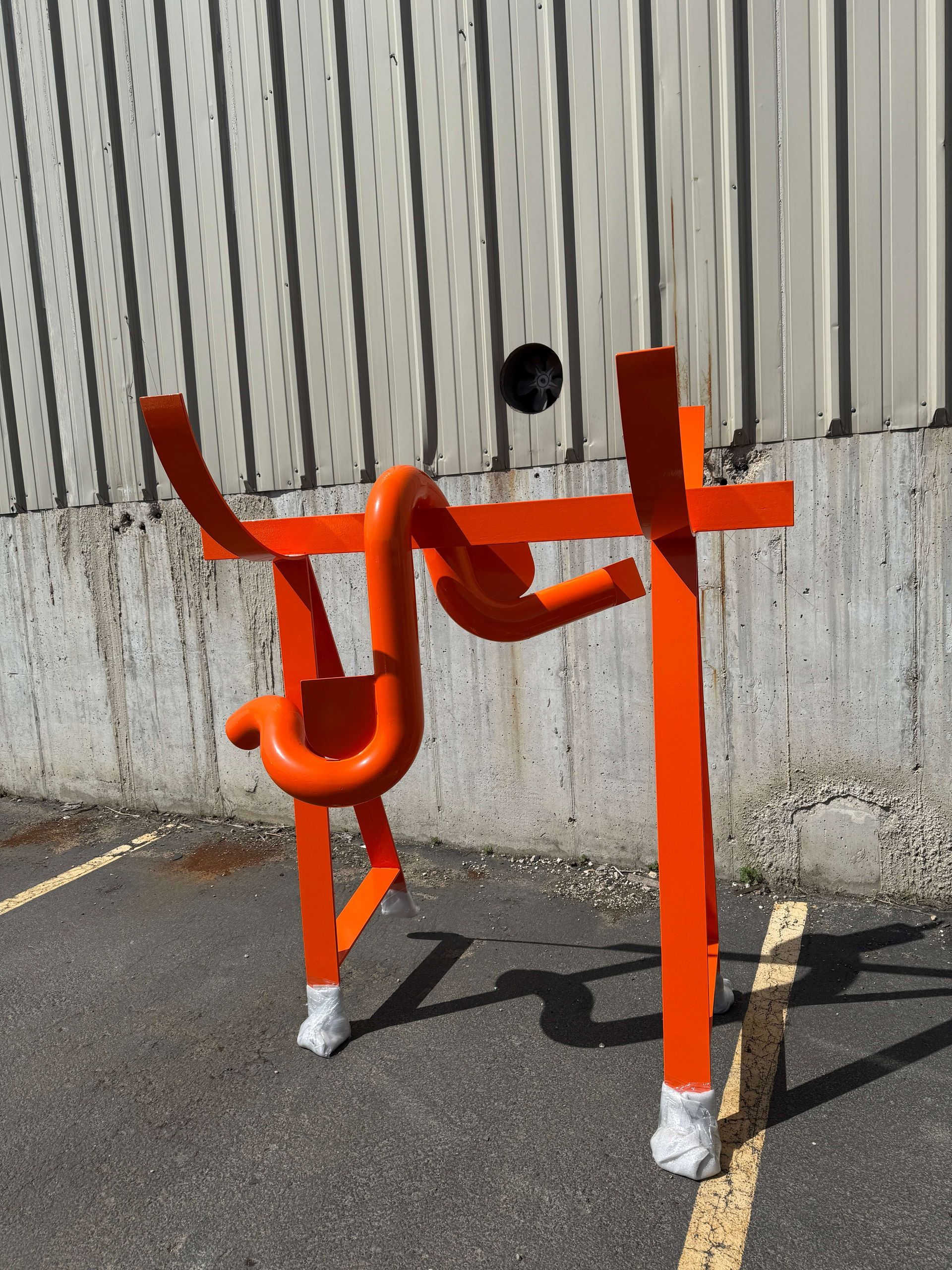 A powder-coated orange sculpture is sitting in a parking lot next to a wall.