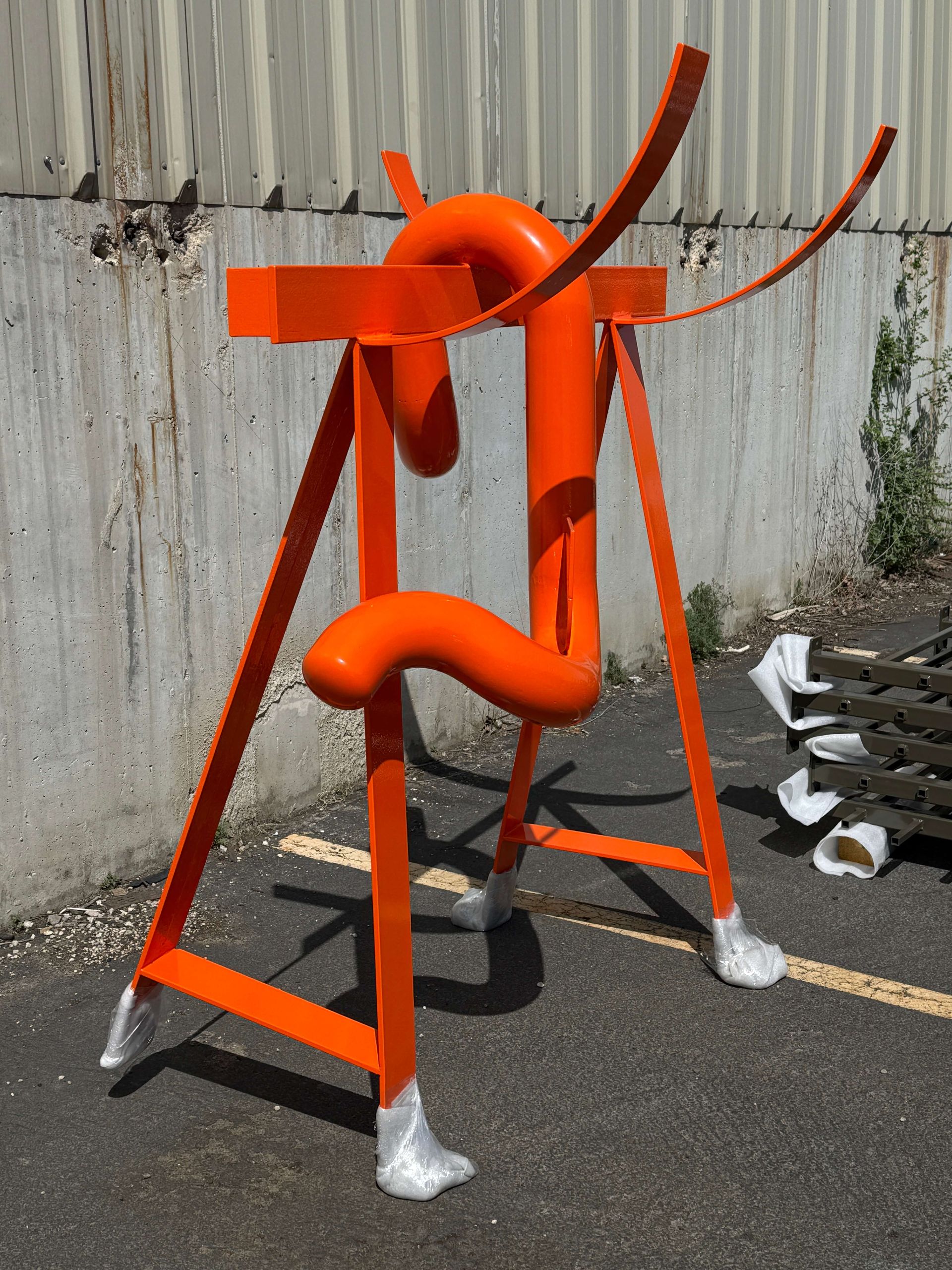 A large orange powder coated sculpture is sitting in a parking lot