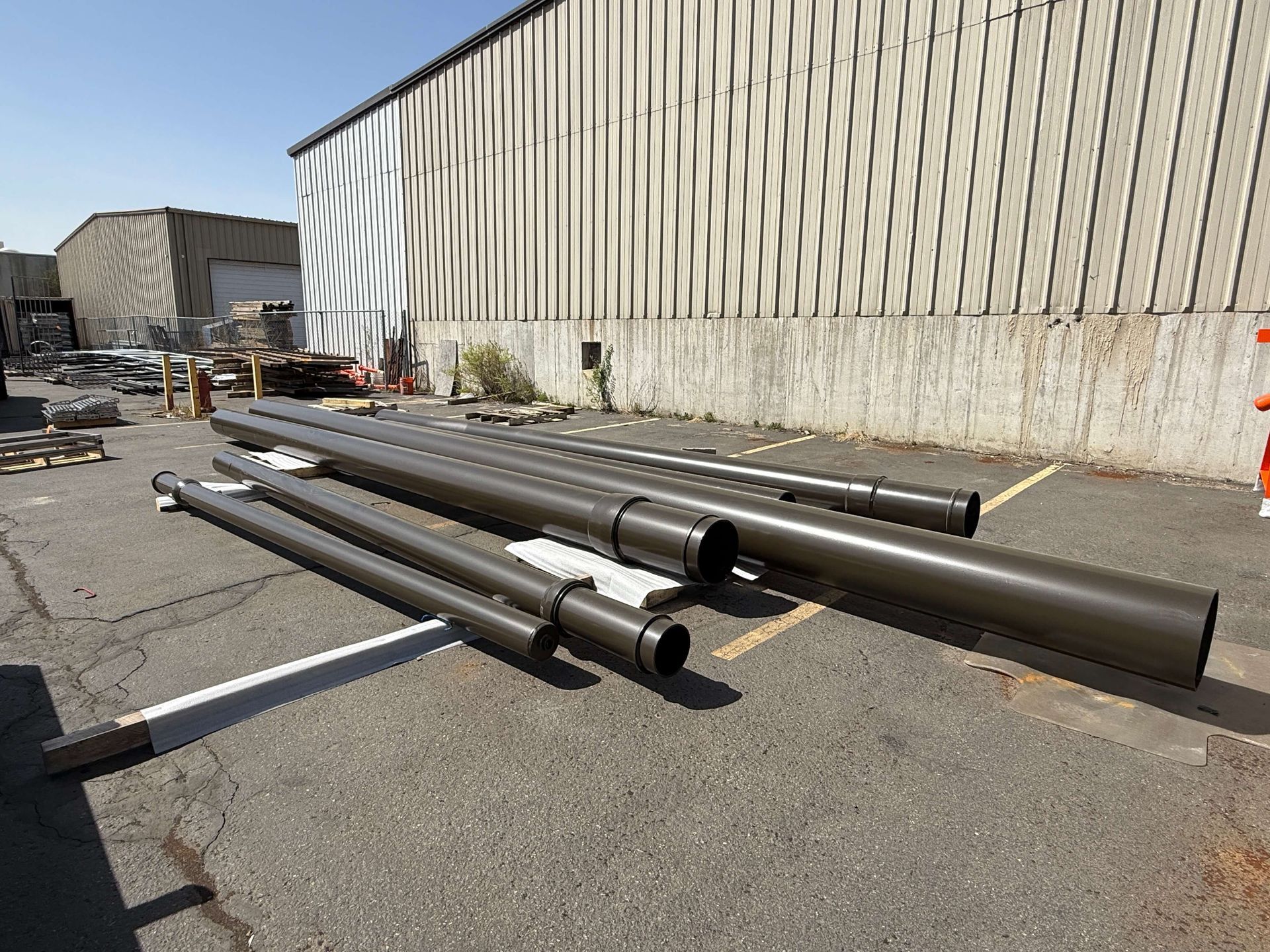 Brown metal poles of varying lengths lying on asphalt in front of a corrugated metal building.
