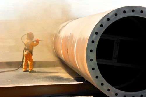 Worker in protective gear sandblasting a large metal pipe.