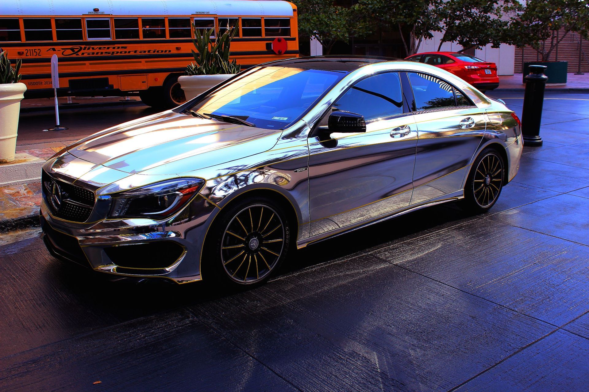 Silver Mercedes sedan parked on a wet street at dusk with an orange bus in the background