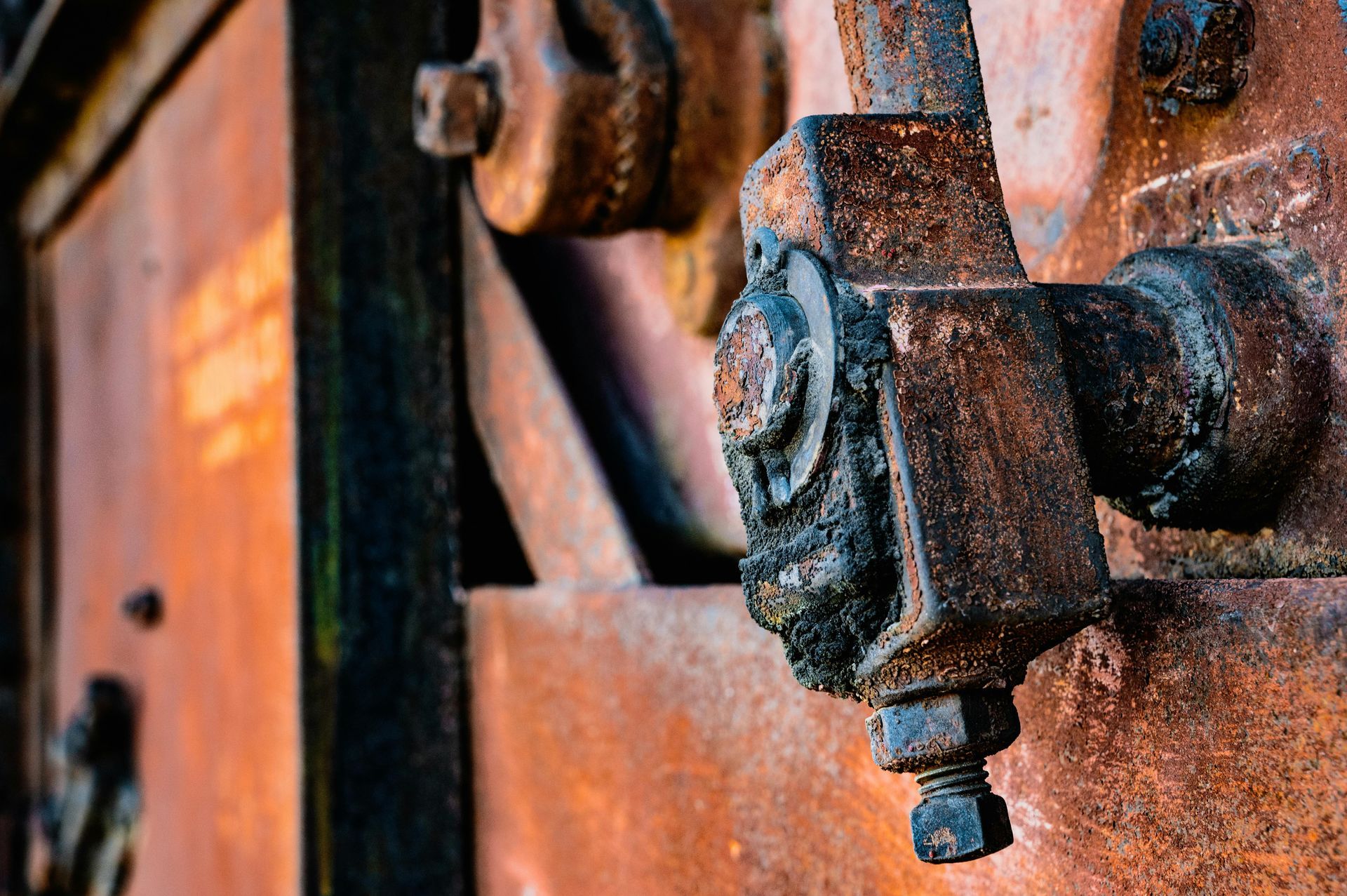 A close-up of a weathered, rusted metal mechanical component with gears and bolts against a textured orange-brown background.
