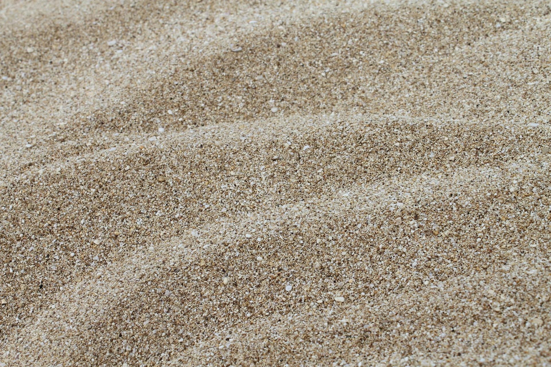 A close-up, high-angle shot of a pile of coarse, light brown sand with gentle ridges running diagonally across the surface.