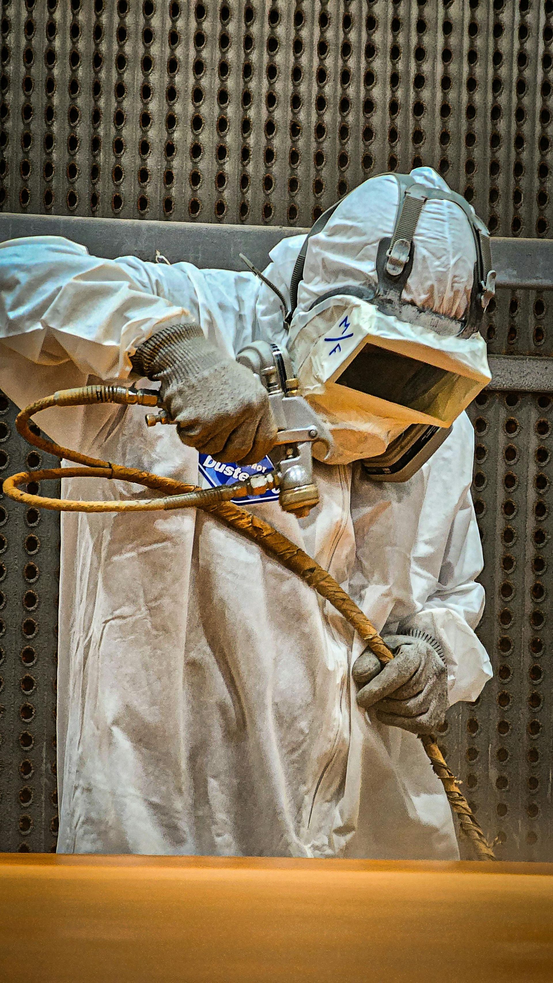 Person in protective suit, sandblasting a surface in an industrial setting.
