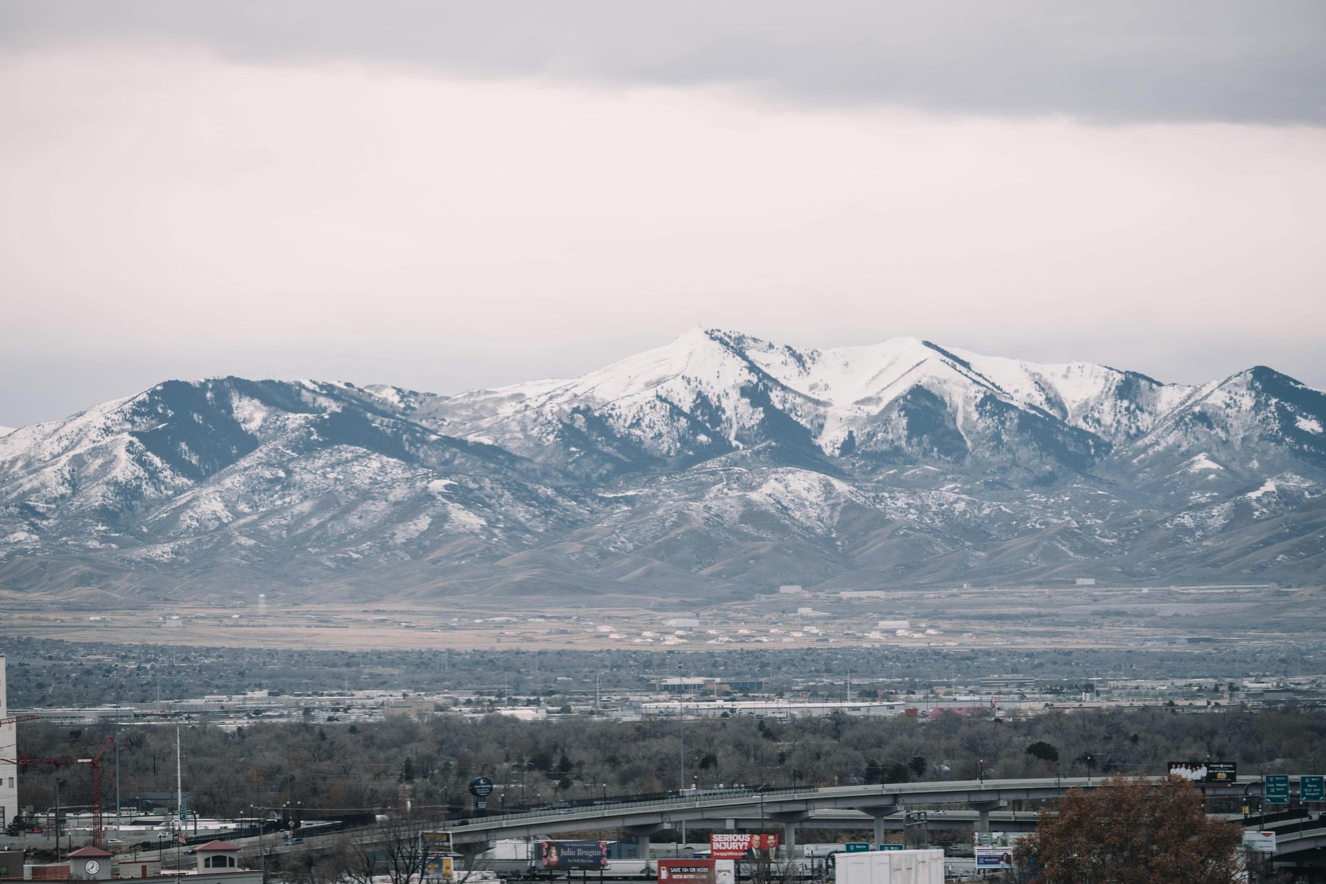 A snowy mountain range rises above a city valley under a soft, overcast sky.