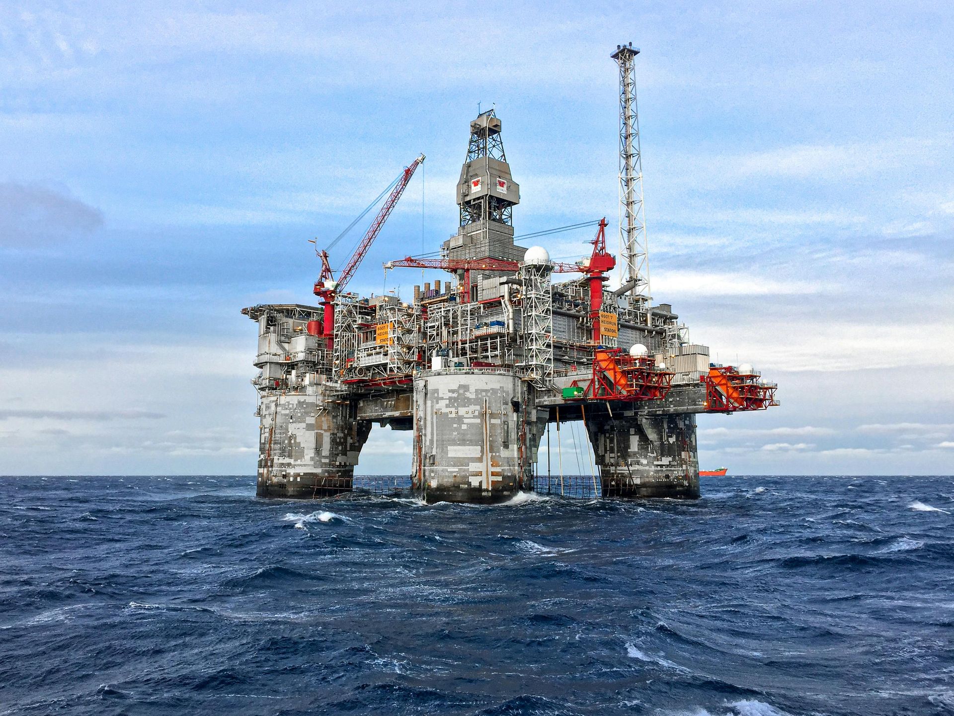 A large, gray oil drilling platform stands in the middle of choppy, dark blue ocean waters under a cloudy sky.