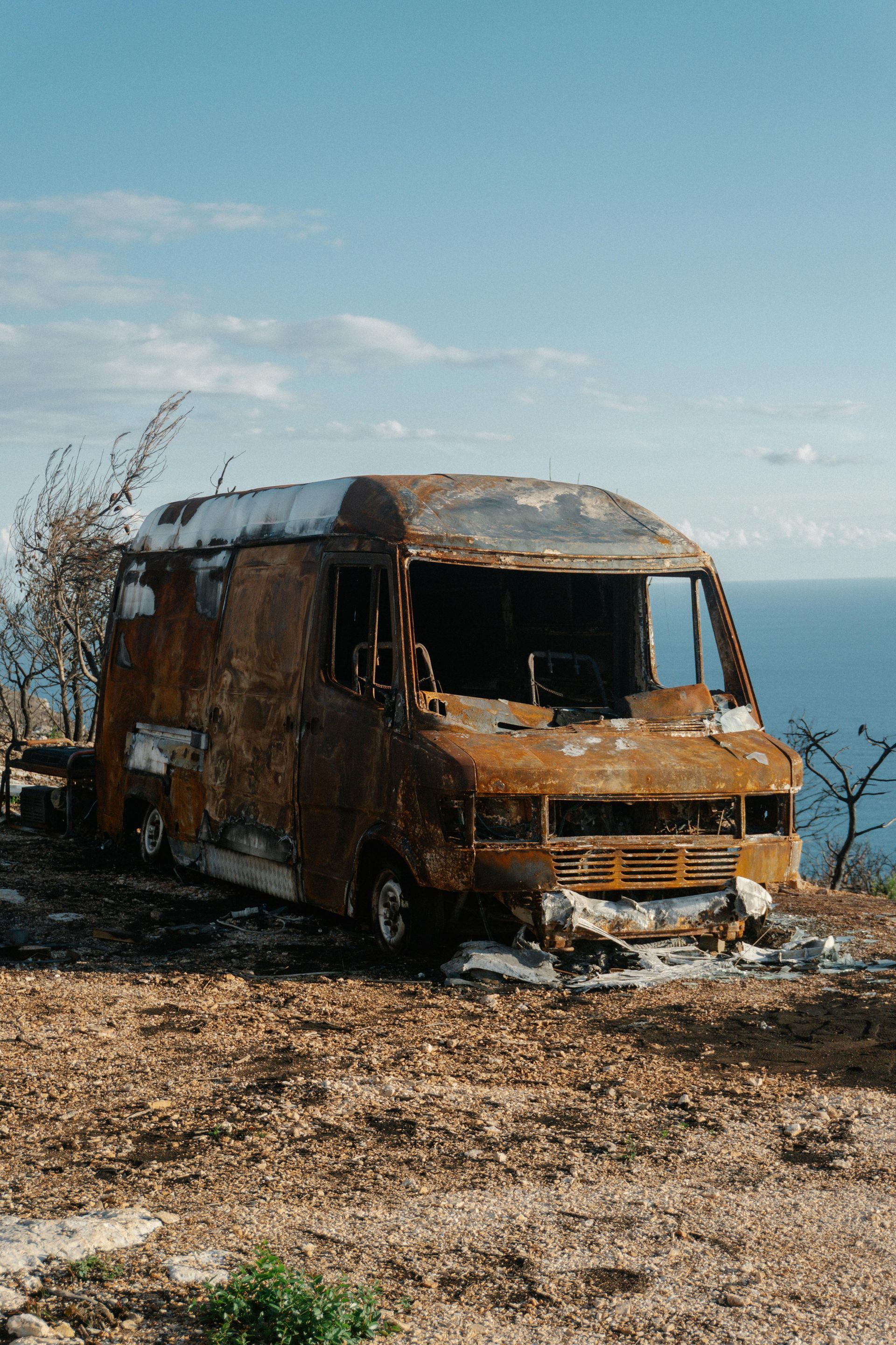 Burnt-out van on a hillside overlooking the ocean under a blue sky; brown and orange hues.
