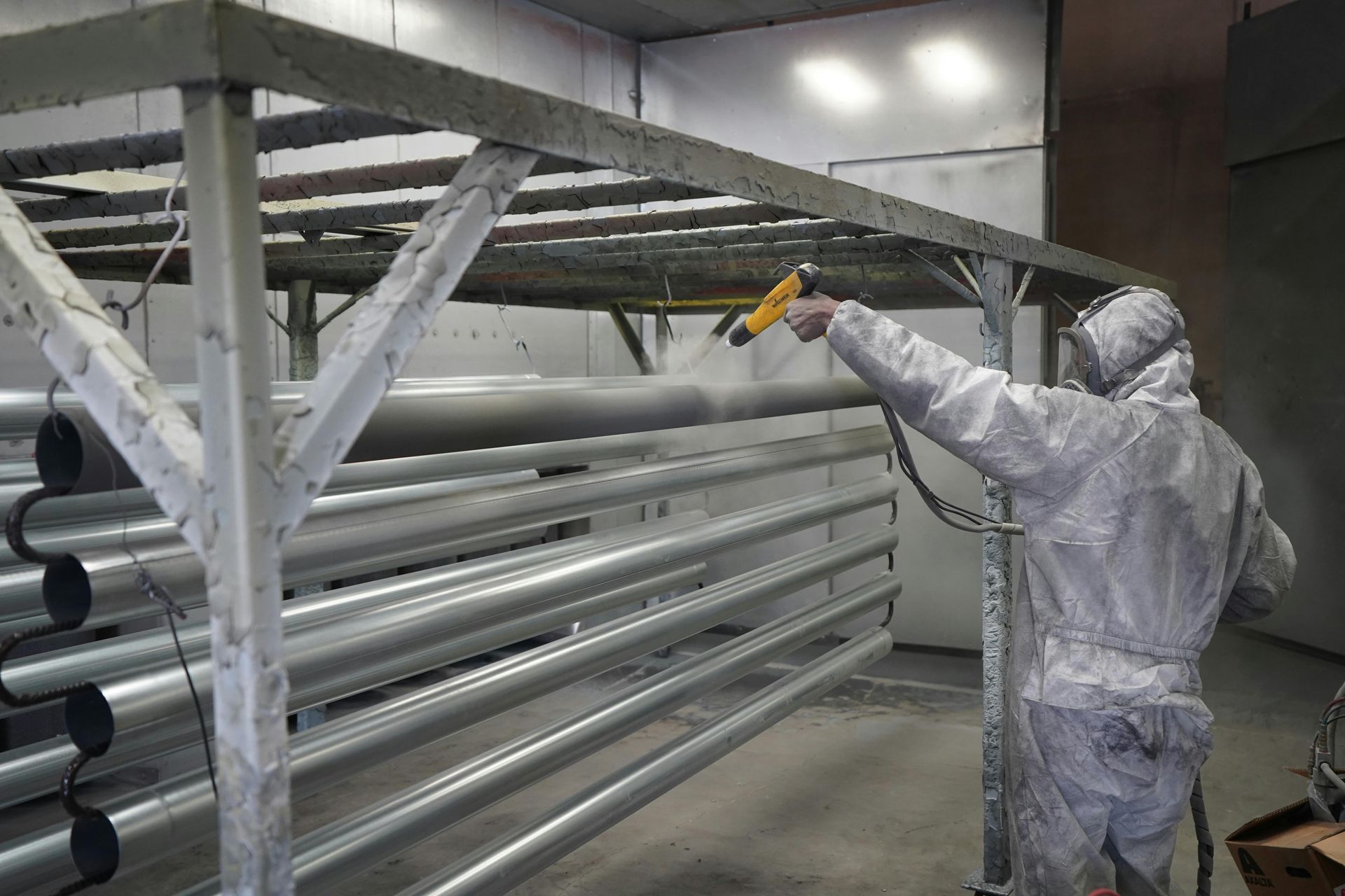 A person in protective coveralls uses a spray gun to apply a powder coating to metal pipes in an industrial booth.