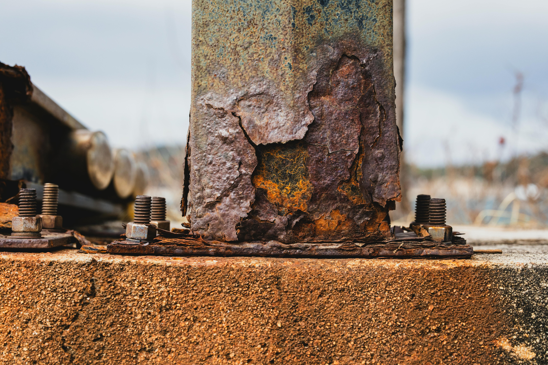 A metal support beam severely corroded with heavy orange rust, bolted to a concrete foundation outdoors.