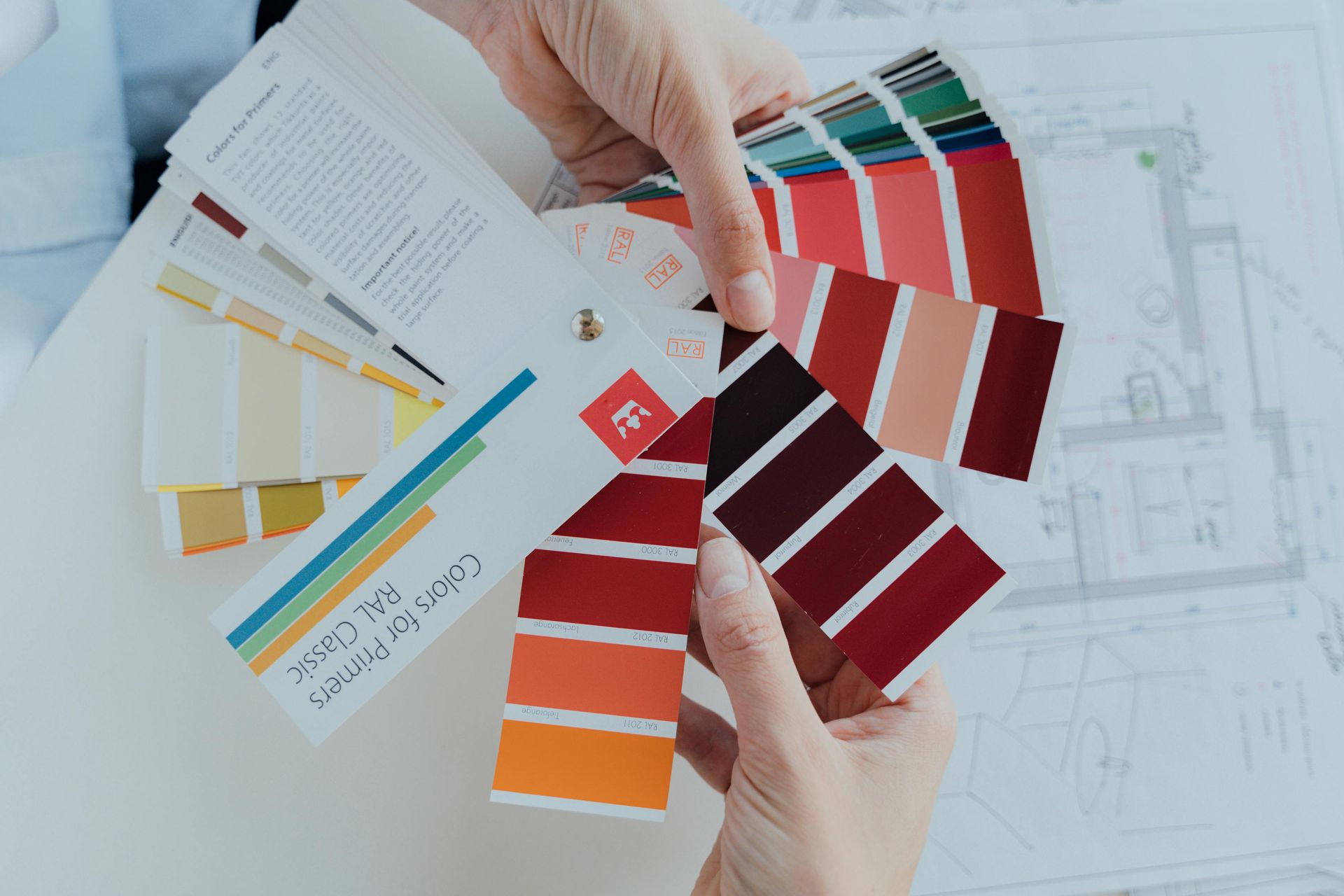 Hands holding color swatch fan over architectural plans, showcasing shades of red, orange, and tan.