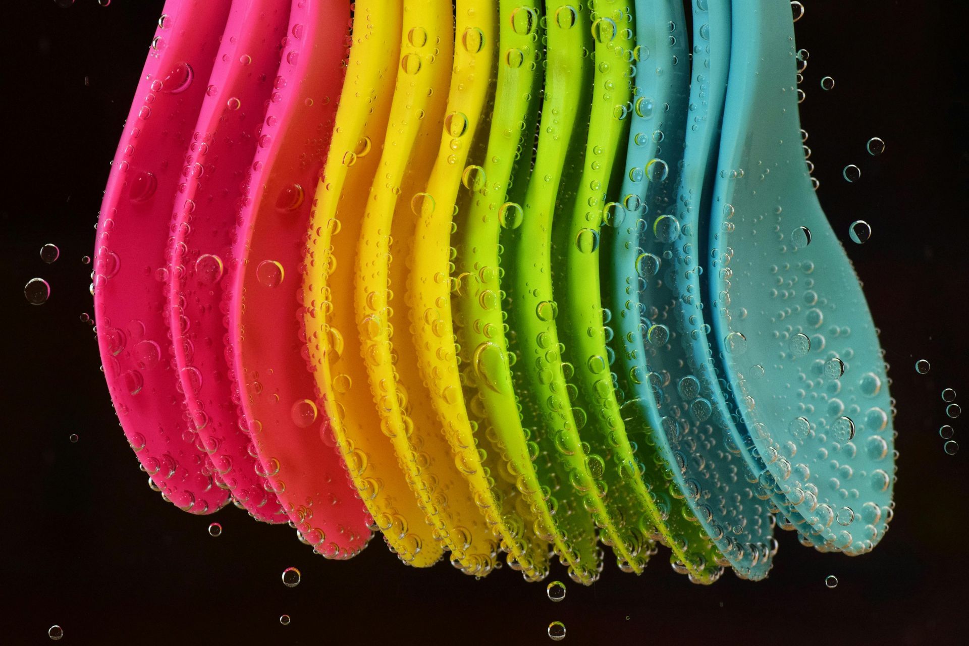 Colorful plastic spoons submerged in water with bubbles, against a black background.