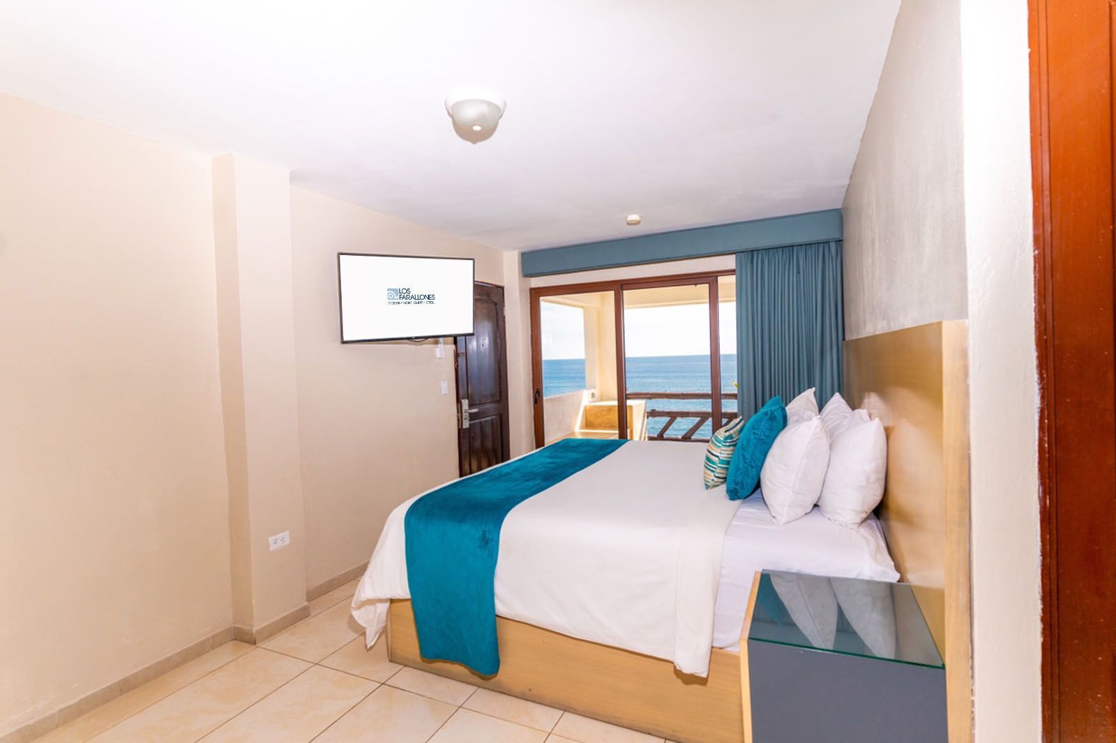 A bedroom with a king-sized bed, teal runner, blue curtains, and a television, featuring an ocean view from a balcony.
