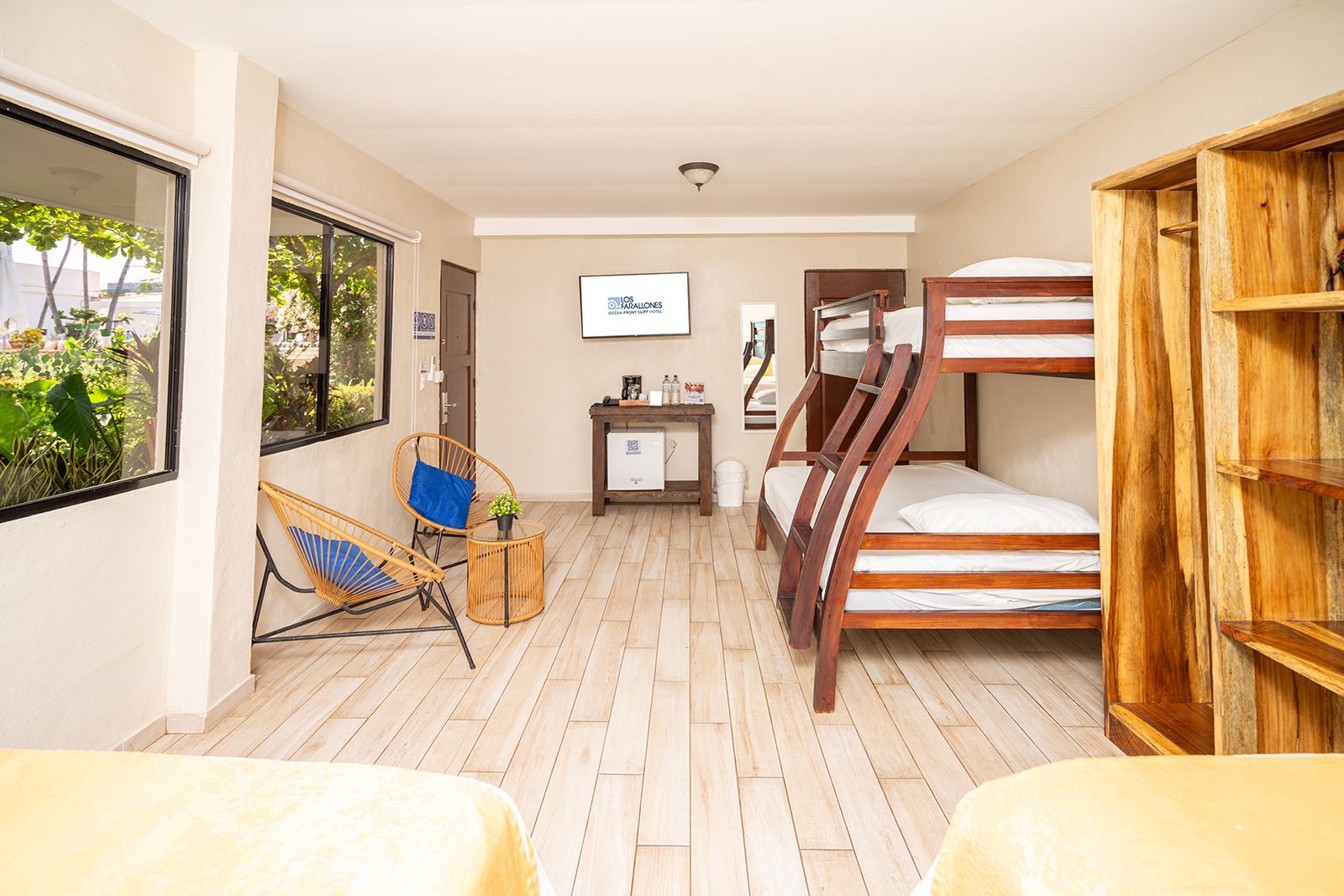 A hotel room with light-colored tile floors, a wooden bunk bed, a small table, chairs, a wall-mounted TV, and a shelf unit.