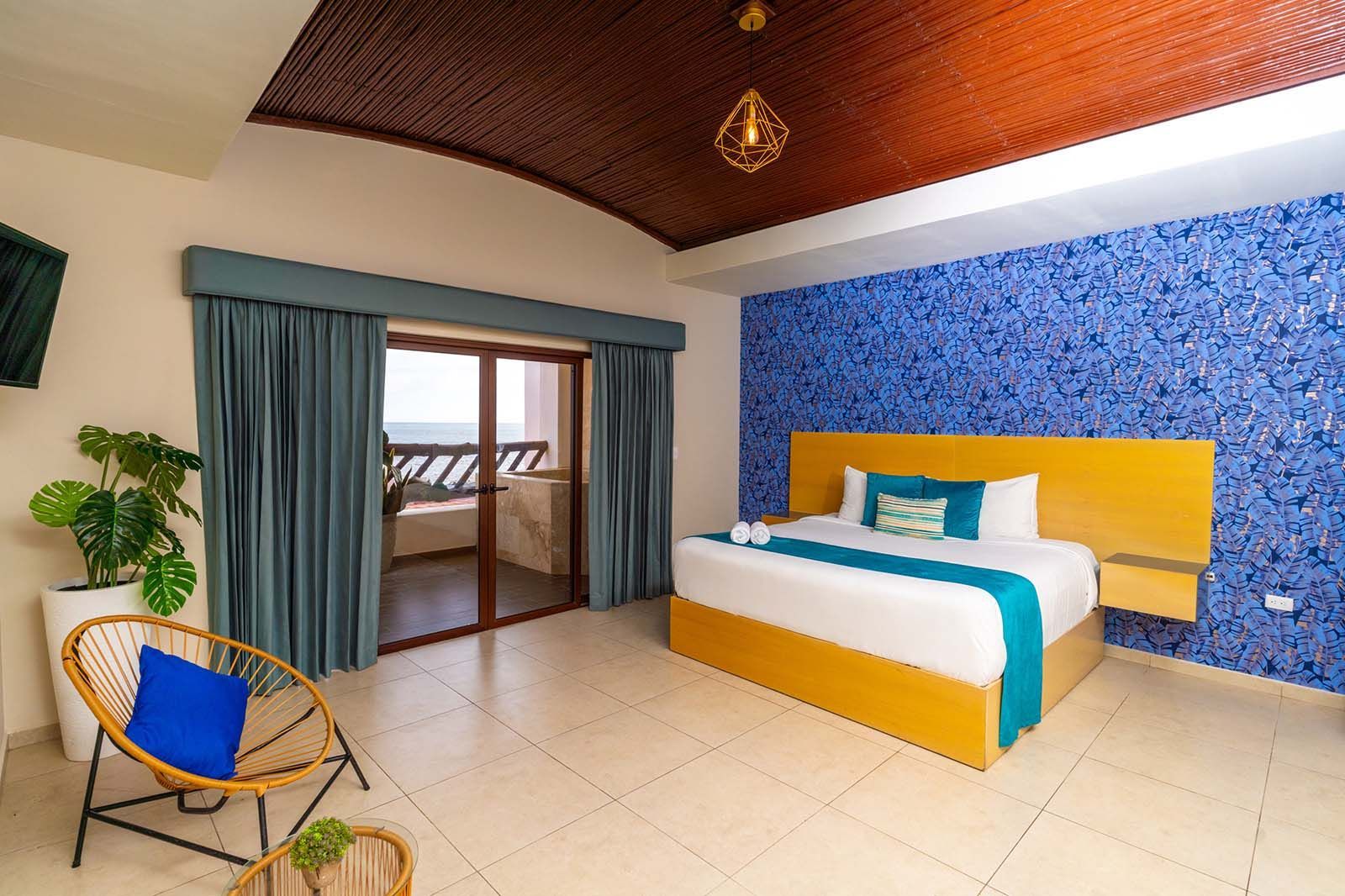 A hotel room with a yellow platform bed, blue patterned accent wall, vaulted ceiling, and a view of the ocean.