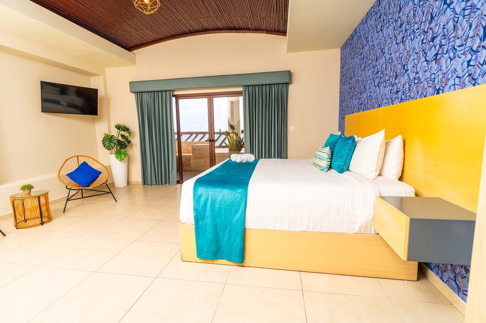 A hotel room with a bed, teal runner, blue accent wall, yellow headboard, and an armchair by a balcony door.