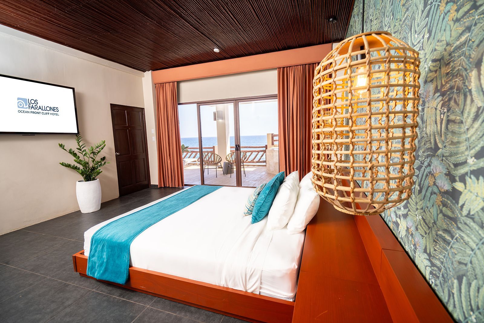 A hotel room with a large bed, a turquoise runner, patterned wallpaper, a hanging wicker lamp, and a view of the sea.