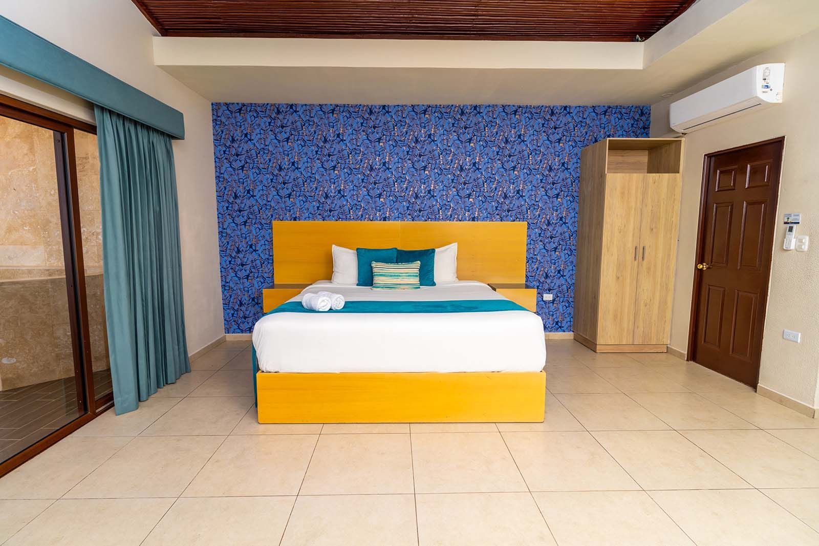 A bedroom with a yellow bed frame, blue accent wall, wooden wardrobe, beige tiled floor, and teal curtains.