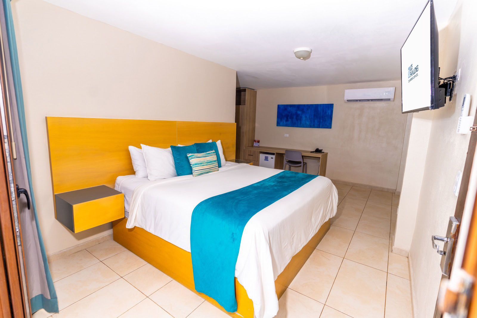 A hotel room featuring a king-size bed with yellow headboard, white linens, and a teal runner, plus a desk and TV.