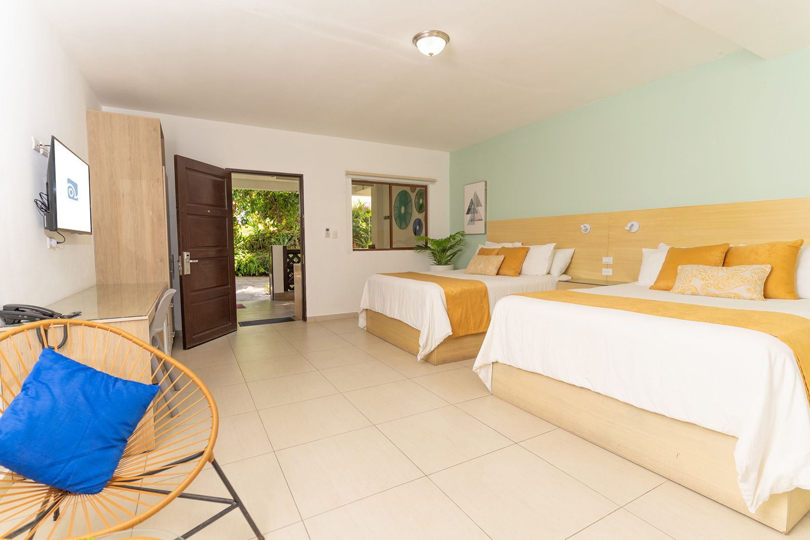 A bright hotel room with two queen beds, light wood headboards, white linens, and a round chair with a blue cushion.
