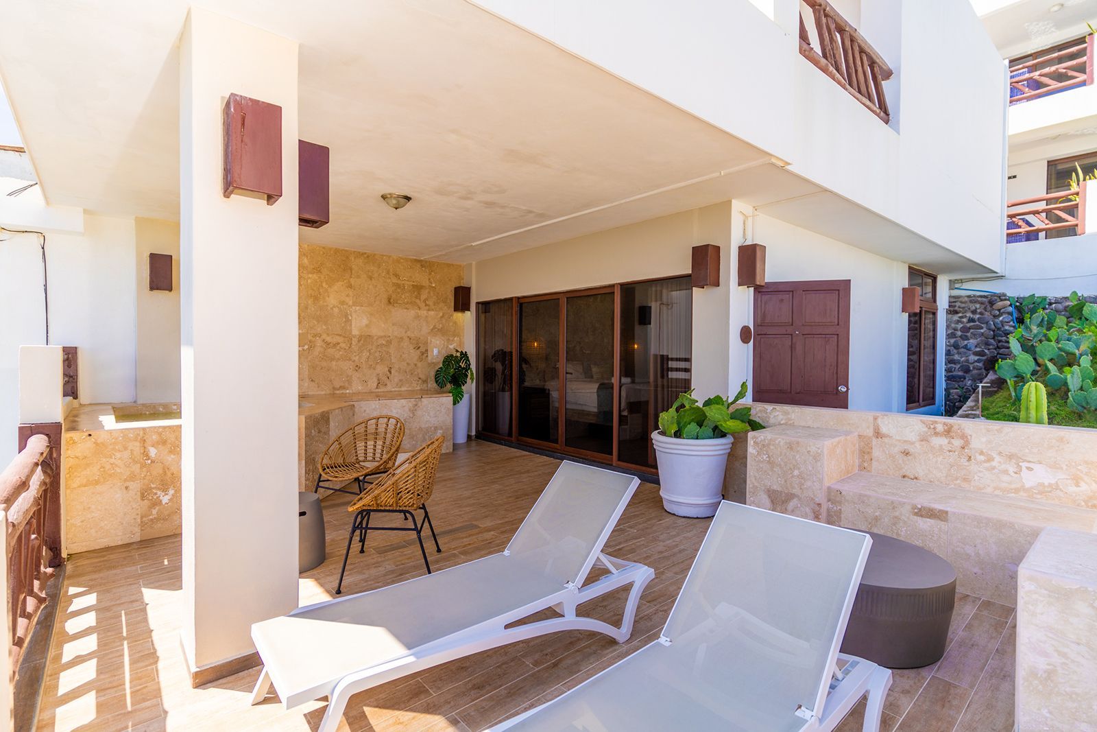 A patio with two white lounge chairs, small woven chairs, and a potted plant in front of a building with stone walls.
