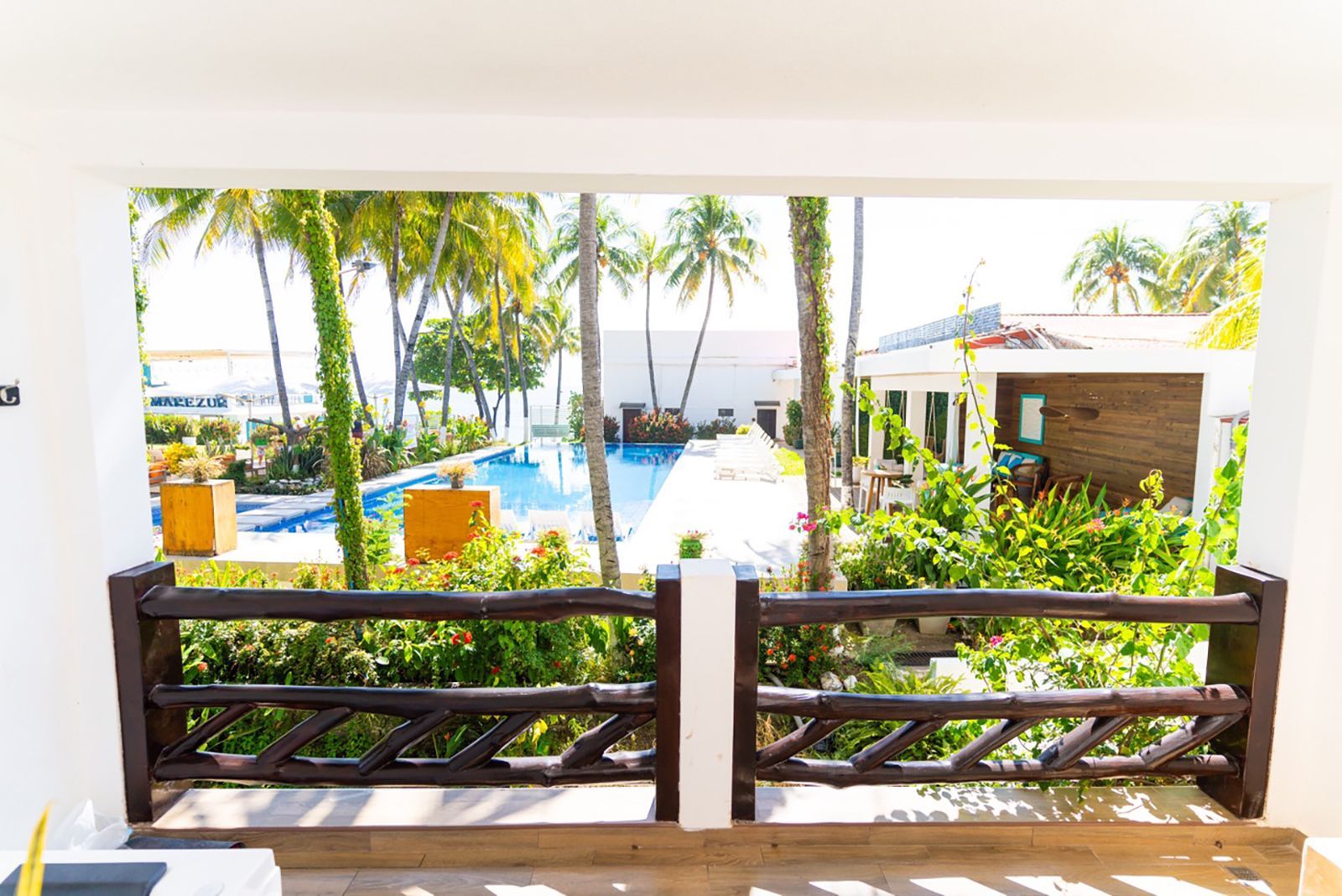 A dark wooden balcony railing overlooks a sunny tropical resort with a blue swimming pool, palm trees, and lush greenery.