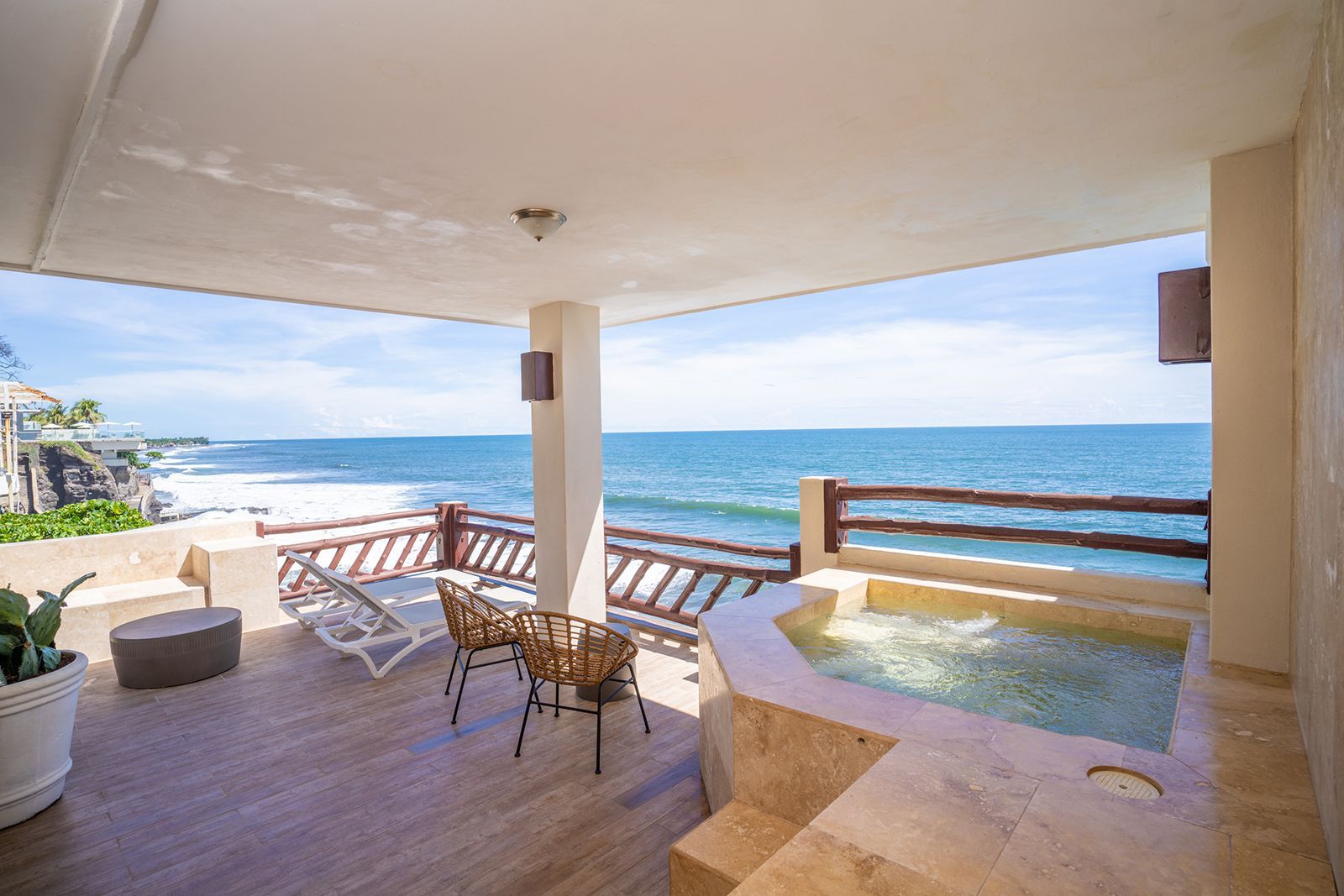 A covered balcony with a hot tub, lounge chairs, and wooden railings overlooking a blue ocean.