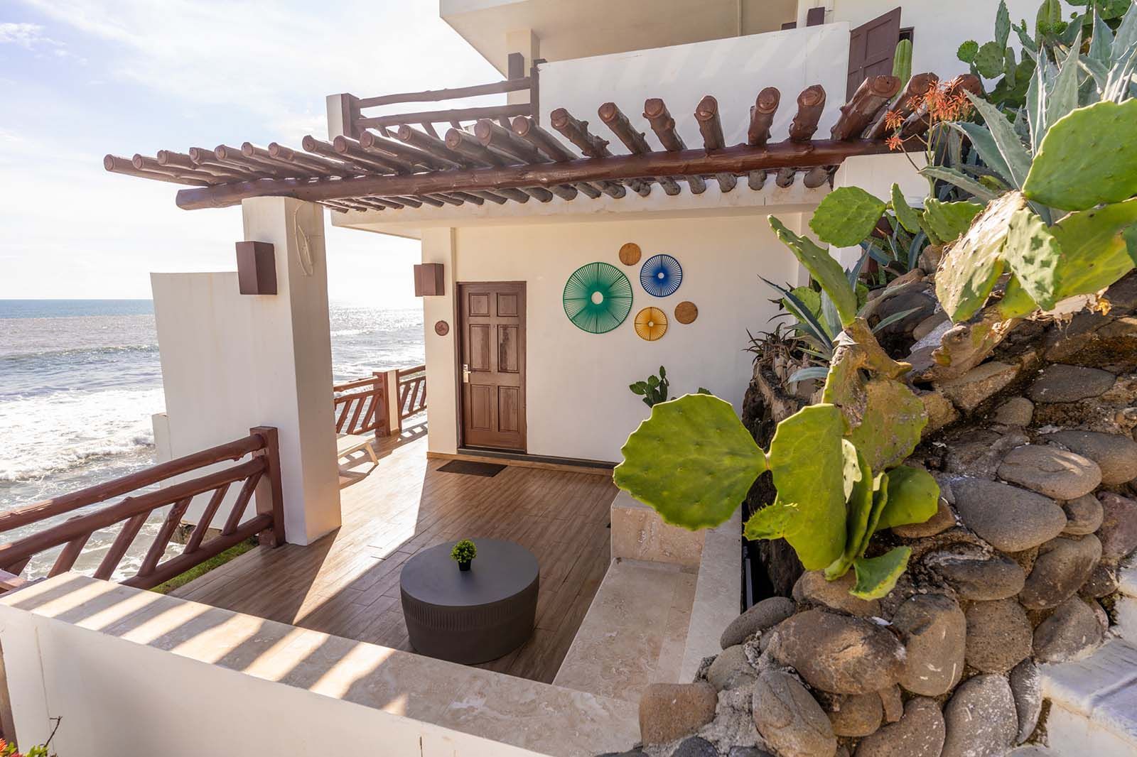 A sunny balcony with a wooden floor, round table, and wooden pergola overlooking the ocean, with cacti and a stone wall.