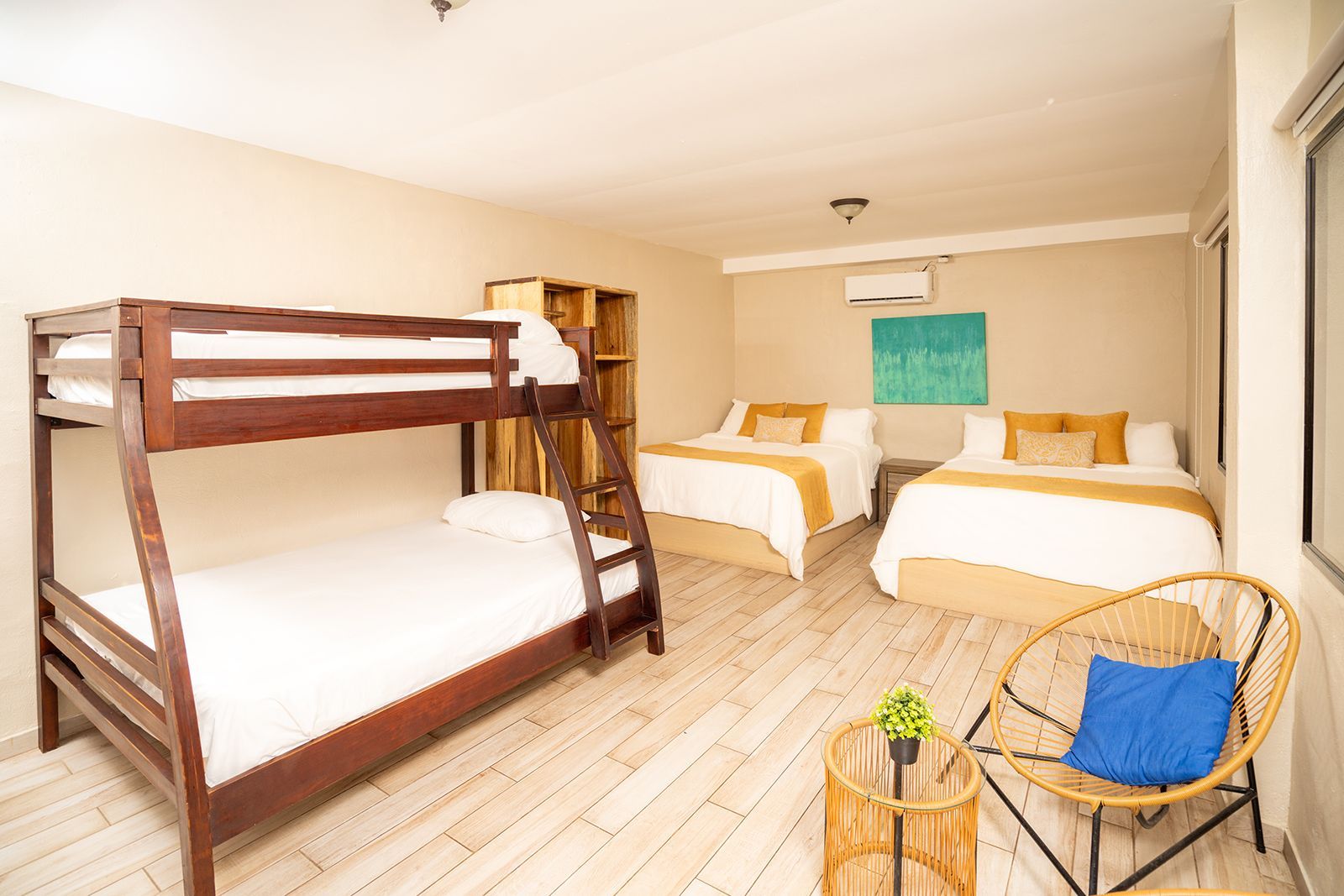 A hotel room with a wooden bunk bed, two queen beds, a small round table with a plant, and a blue-cushioned chair.