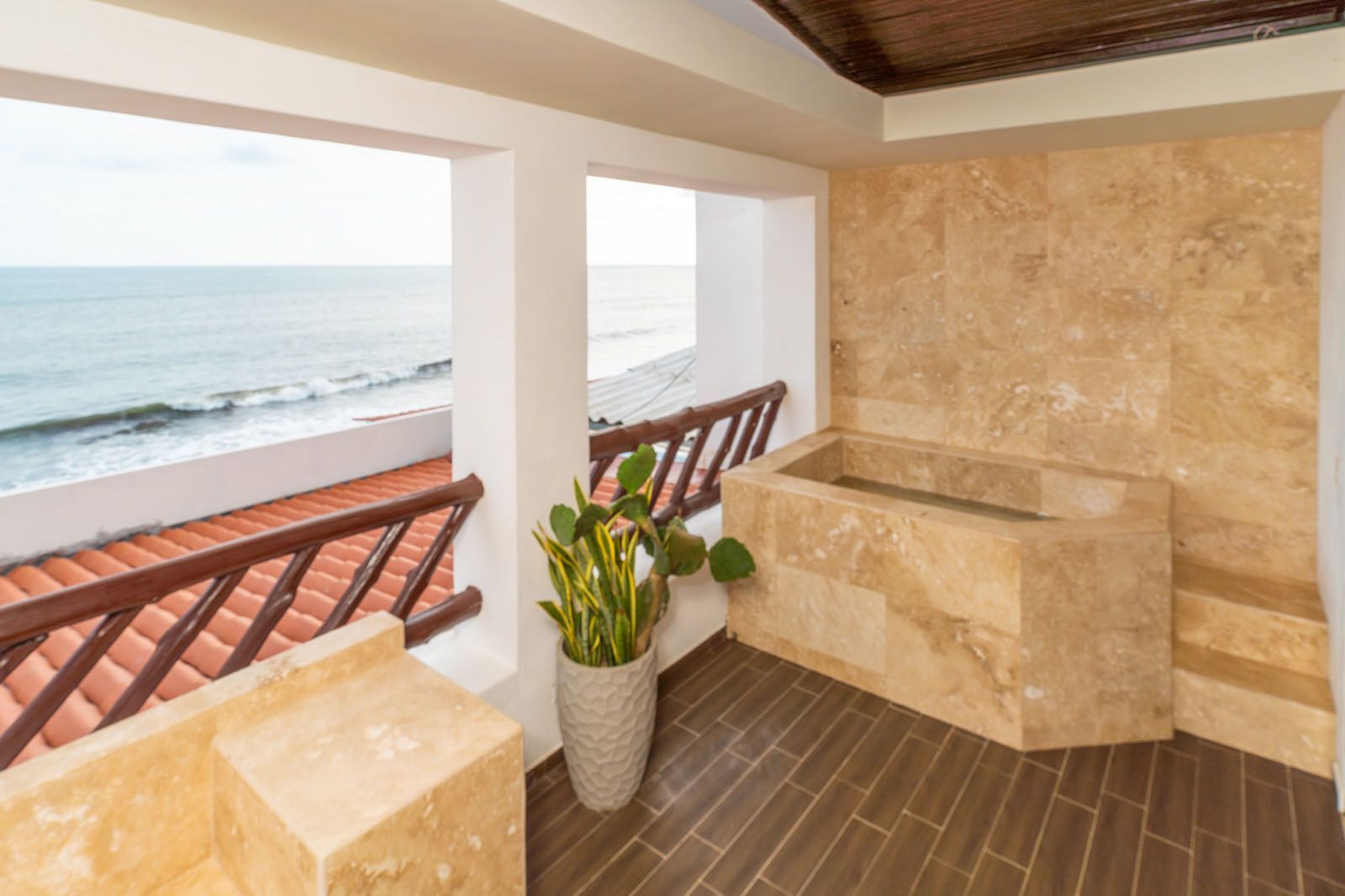 A stone soaking tub on a balcony overlooking the ocean, featuring a potted plant and terracotta-tiled roof below.