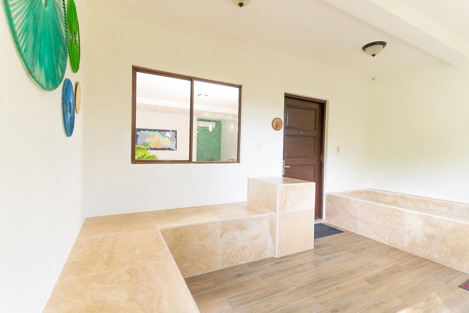 A light-colored room features a stone seating area, a large window, a dark wooden door, and decorative wall plates.