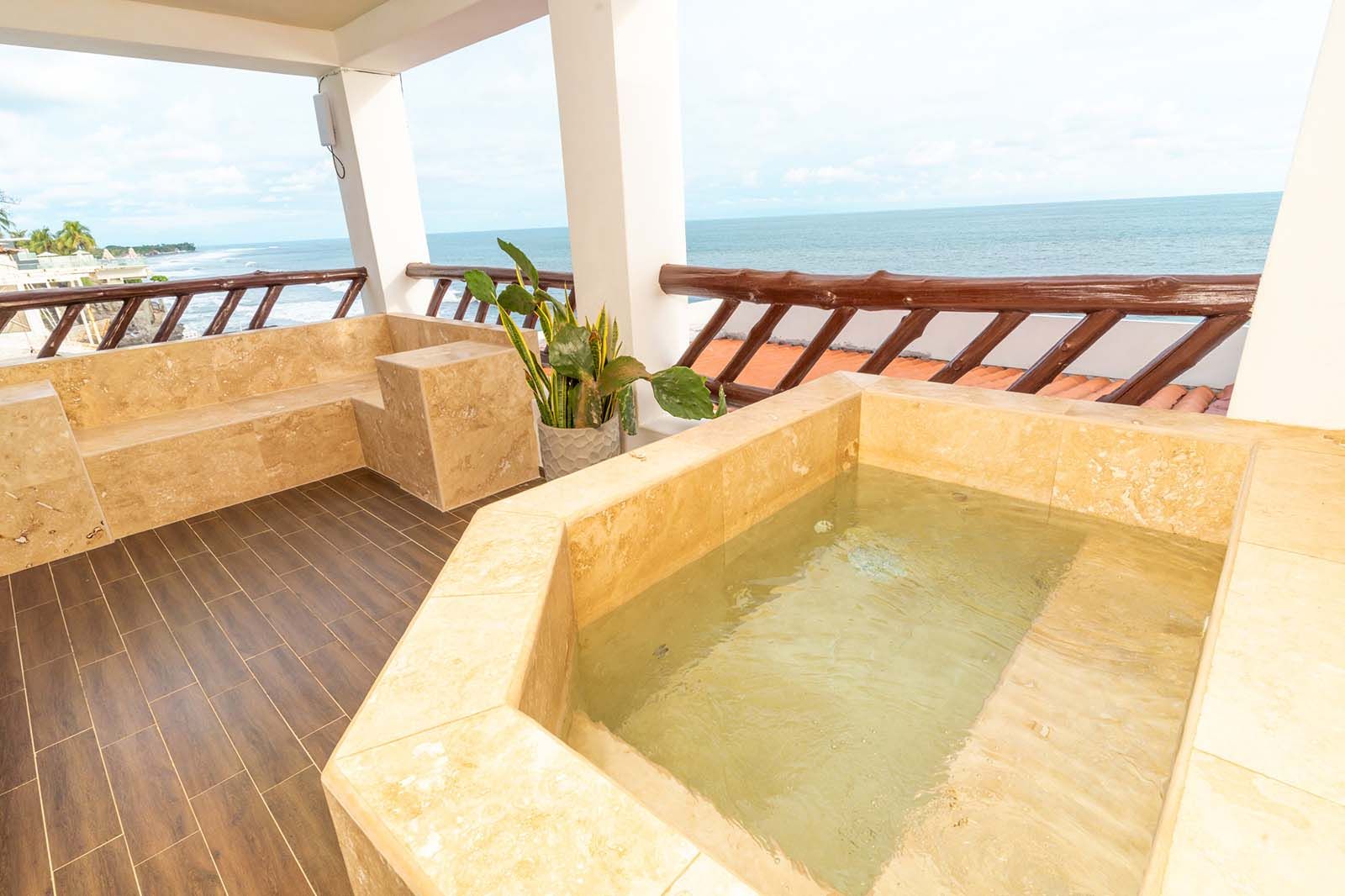 A stone hot tub on a balcony overlooking the ocean with wooden railings and seating.