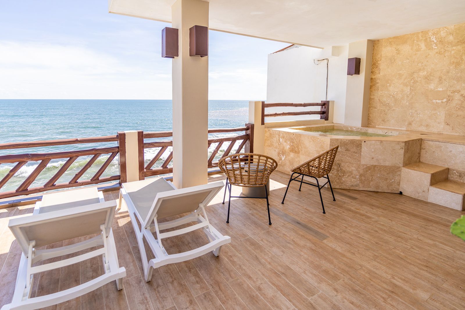 A balcony with two lounge chairs, two chairs, and a small hot tub, overlooking the ocean under a bright sky.