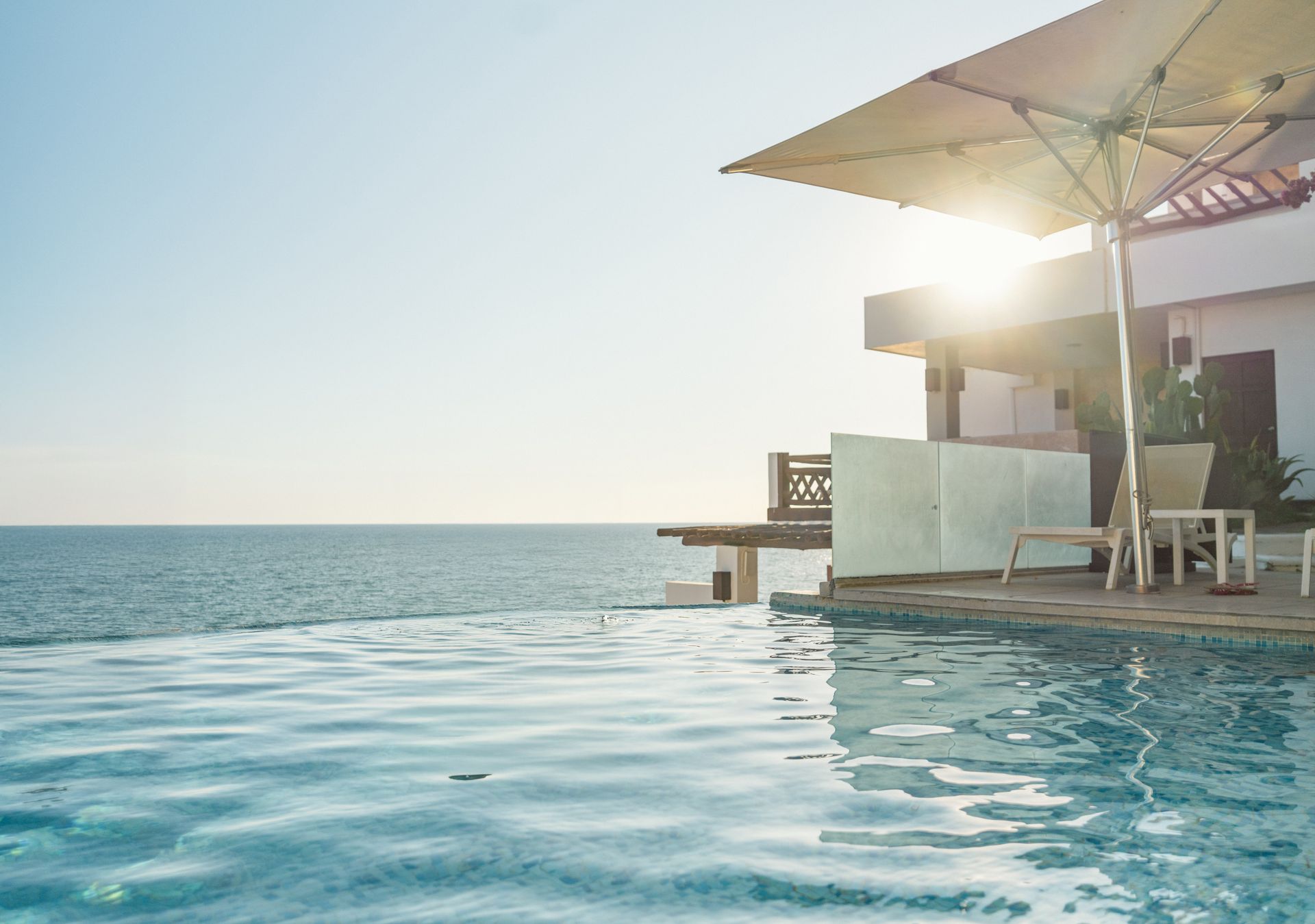 An infinity pool overlooking the ocean at sunset with a lounge chair and umbrella on the patio.