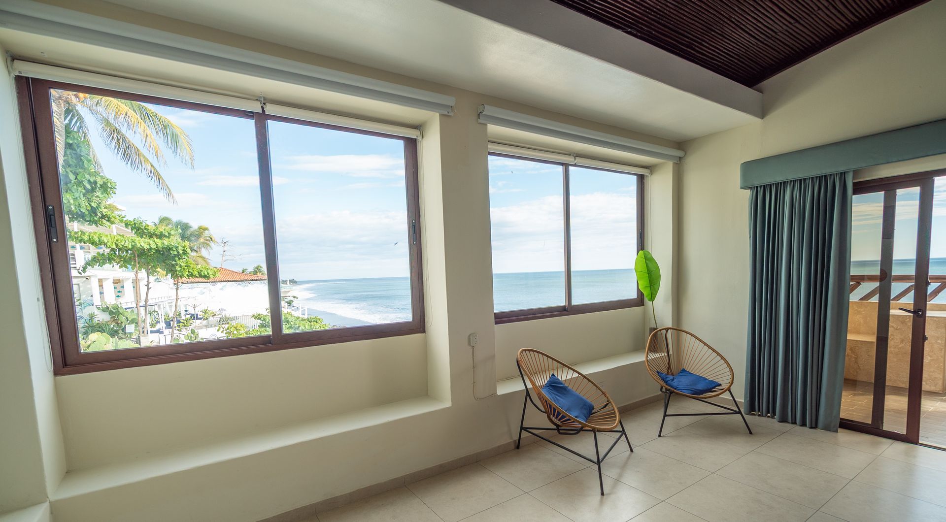 Two rattan chairs in a sunlit room facing floor-to-ceiling windows with a scenic ocean and beach view.