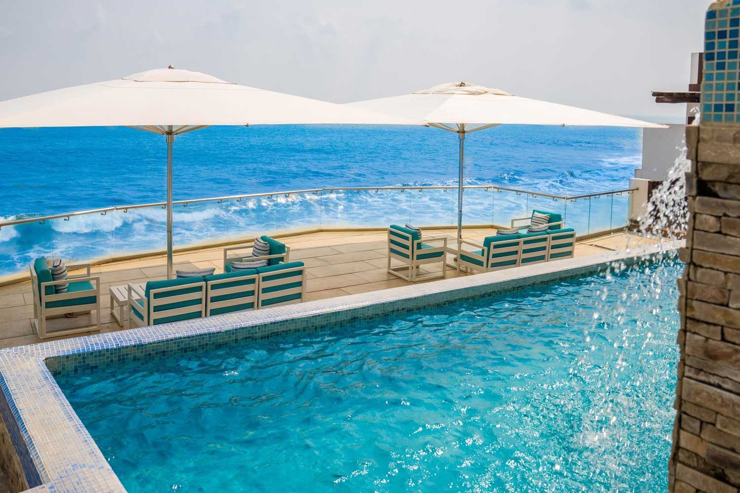 A swimming pool with a waterfall feature overlooks the ocean, with outdoor seating under white umbrellas on a patio.