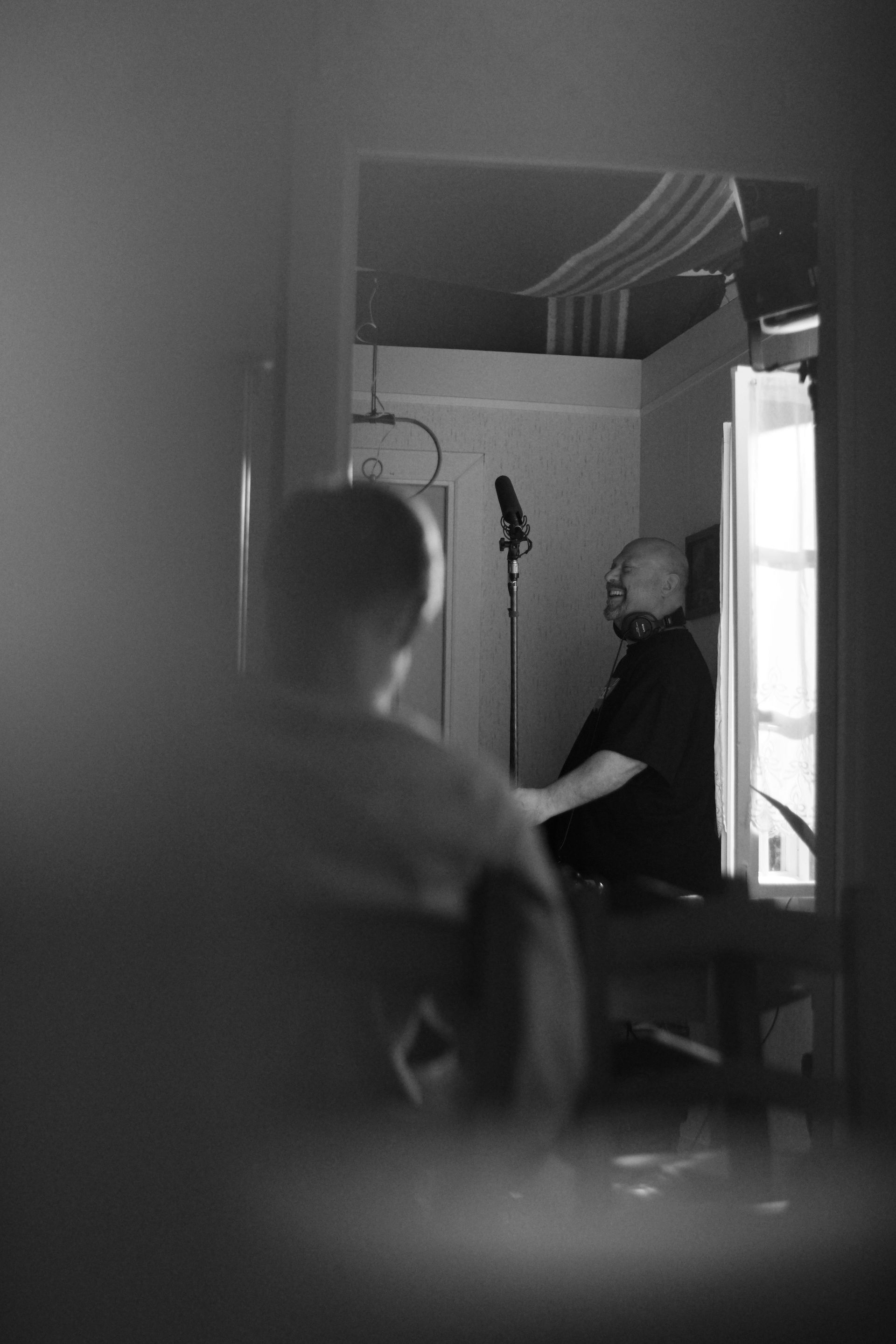 Op deze zwart-witfoto zit een persoon voor een microfoon in een kamer, van achteren gezien door een deuropening.
