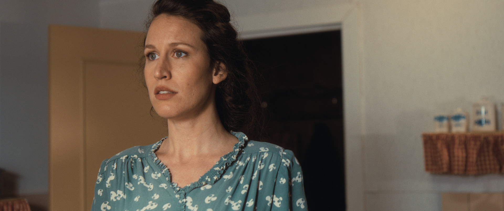 Een persoon met golvend bruin haar, gekleed in een turquoise blouse met bloemenprint, kijkt met een verraste uitdrukking omhoog in een lichte ruimte.