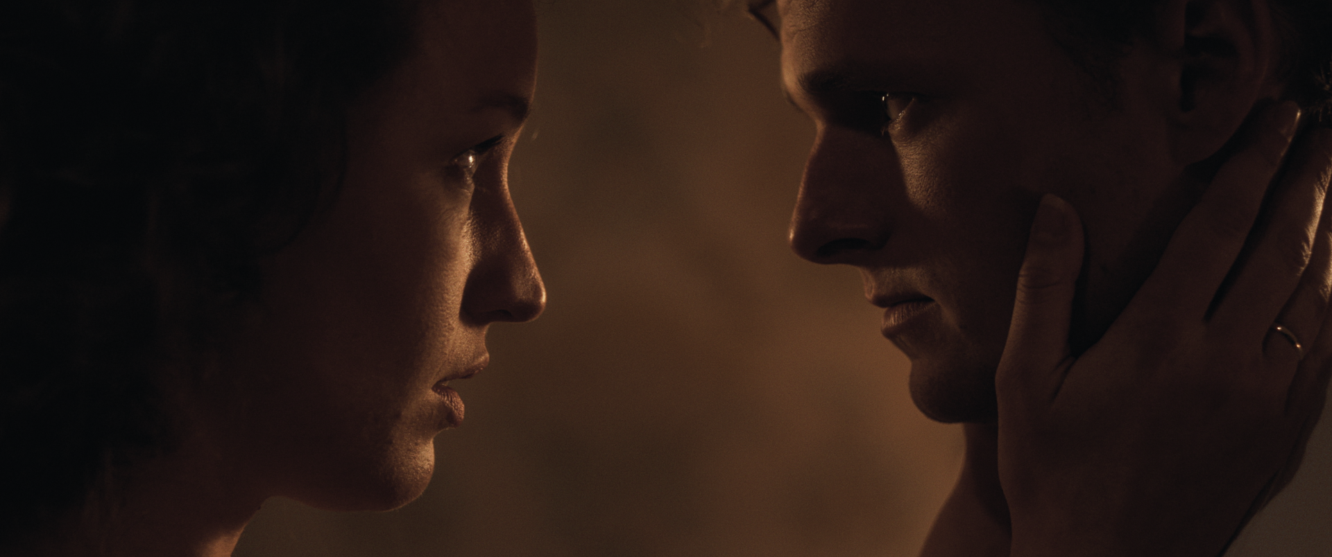 Een close-up van twee mensen die tegenover elkaar staan in gedempt licht, waarbij de ene persoon zachtjes de wang van de ander vasthoudt.