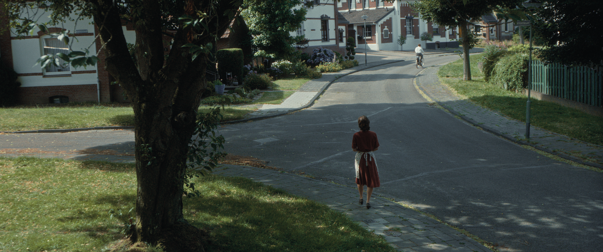 Een vrouw in een rode jurk loopt door een rustige straat in een buitenwijk, terwijl in de verte een fietser nadert.
