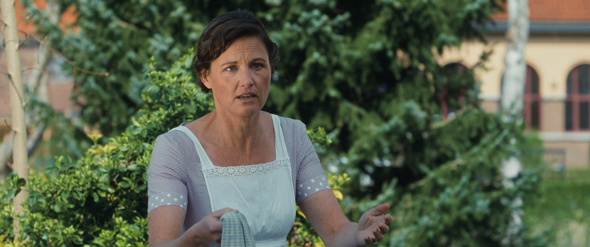 Een persoon met kort donker haar en een lichtgekleurde jurk gebaart terwijl hij spreekt in een tuin.