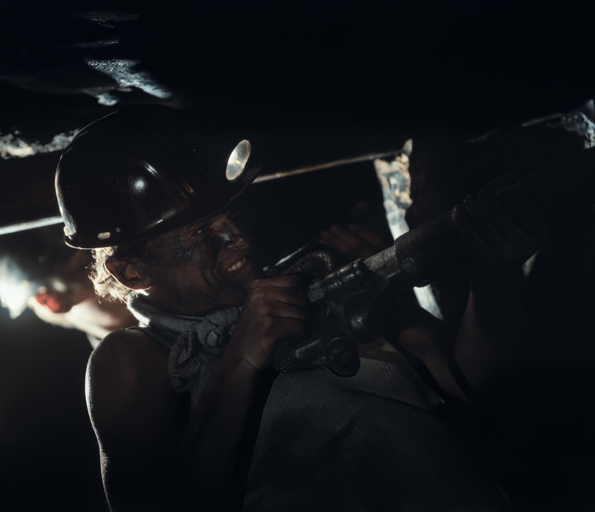 Een arbeider met een veiligheidshelm en hoofdlamp bedient een handboormachine in een slecht verlichte, smalle ondergrondse mijntunnel.