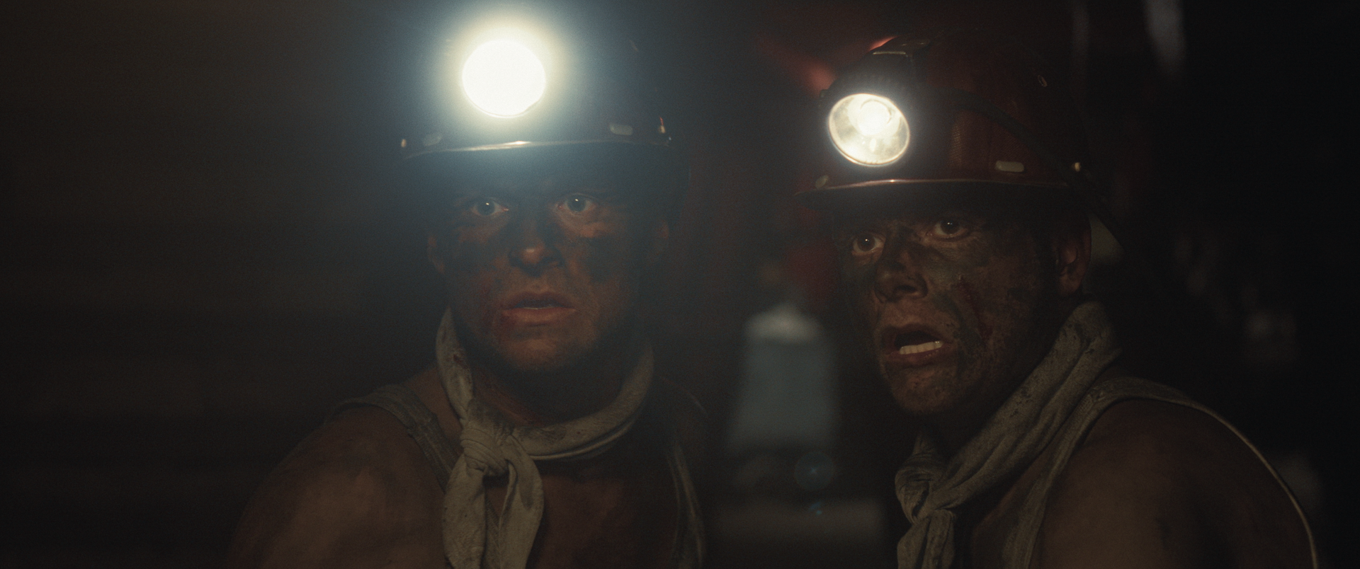 Twee mijnwerkers met roetbevlekte gezichten en brandende koplampen op hun helmen kijken verbaasd voor zich uit.