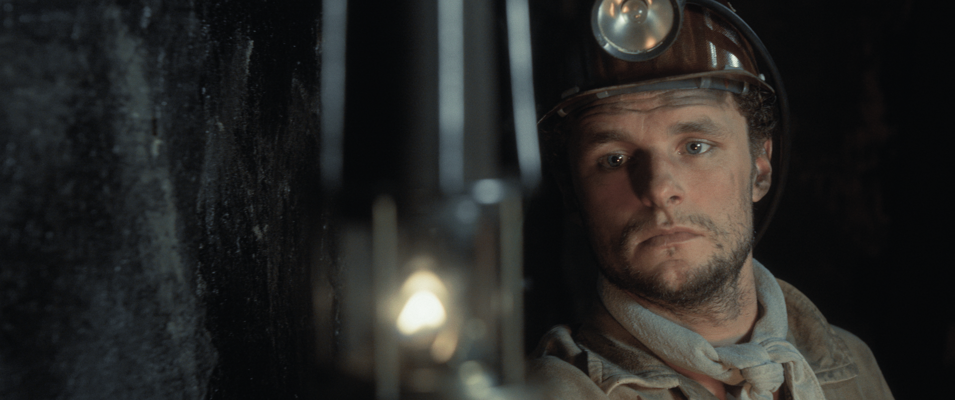 Een mijnwerker met een helm en een hoofdlamp staart intens naar een klein, gloeiend lichtje in een donkere mijn.