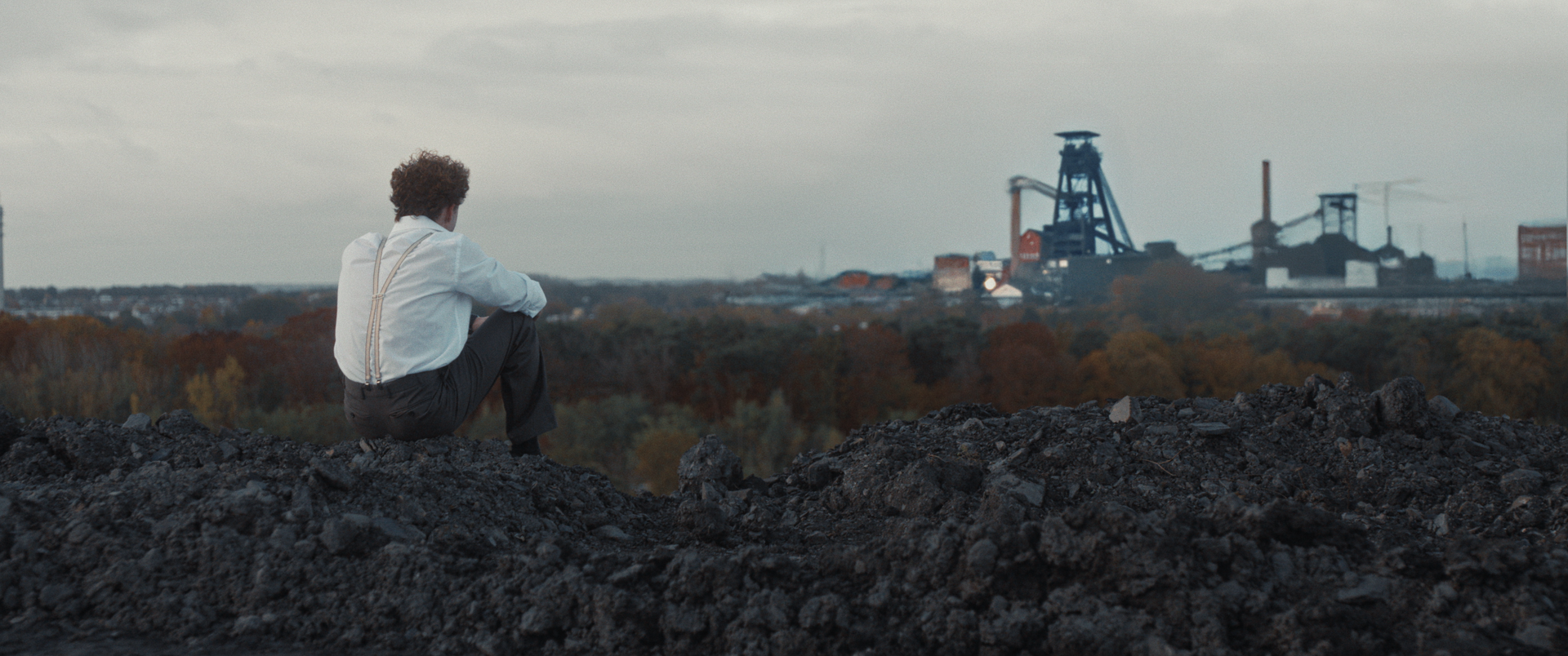 Een persoon in een wit overhemd en bretels zit op een donkere, rotsachtige heuvel en kijkt uit over een industriële fabriek in de verte.